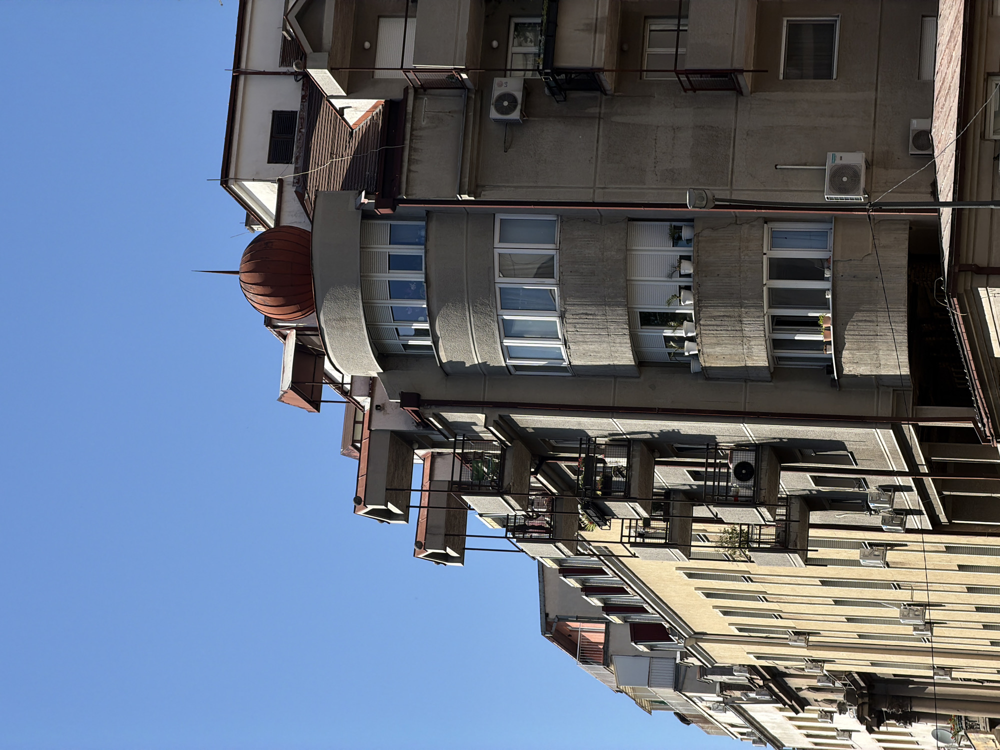The image shows a low-angle view of buildings in Belgrade, Serbia. The buildings are varied in architectural style. One building features a prominent red dome with a spire. Facades show a mix of concrete and painted surfaces. Some windows have air conditioning units mounted below. Small balconies are visible on one of the buildings. The sky is clear and blue.