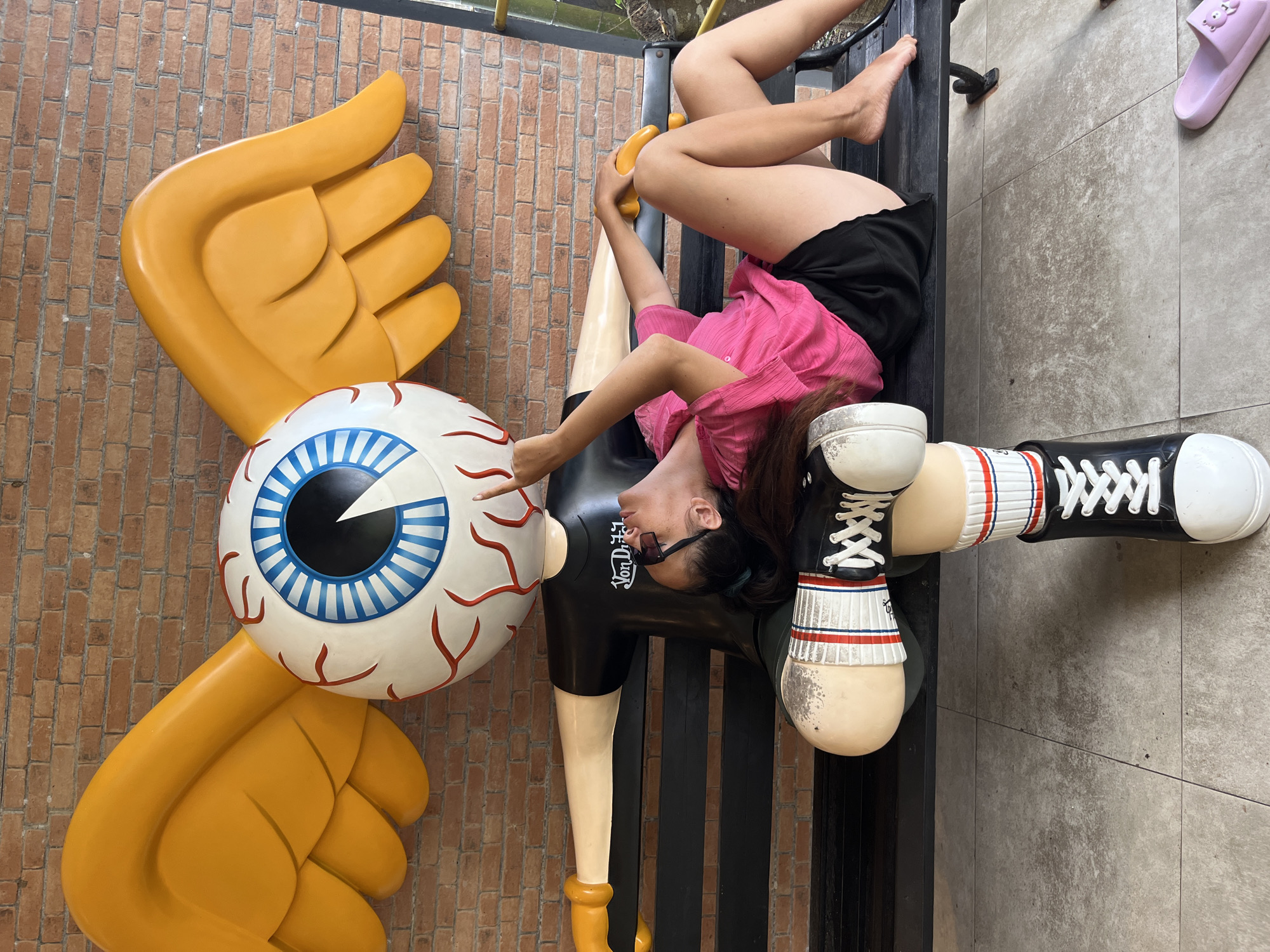 A woman is lying on a black bench next to a large, cartoonish statue of a winged eyeball wearing a black t-shirt with a "Von Dutch" logo. The woman is wearing a pink shirt and black shorts, with sunglasses on her face. The statue has large, yellow wings and is positioned as if lying down as well. The background is a brick wall. The scene appears to be outdoors, possibly in a public area or a themed establishment, given the unusual statue.