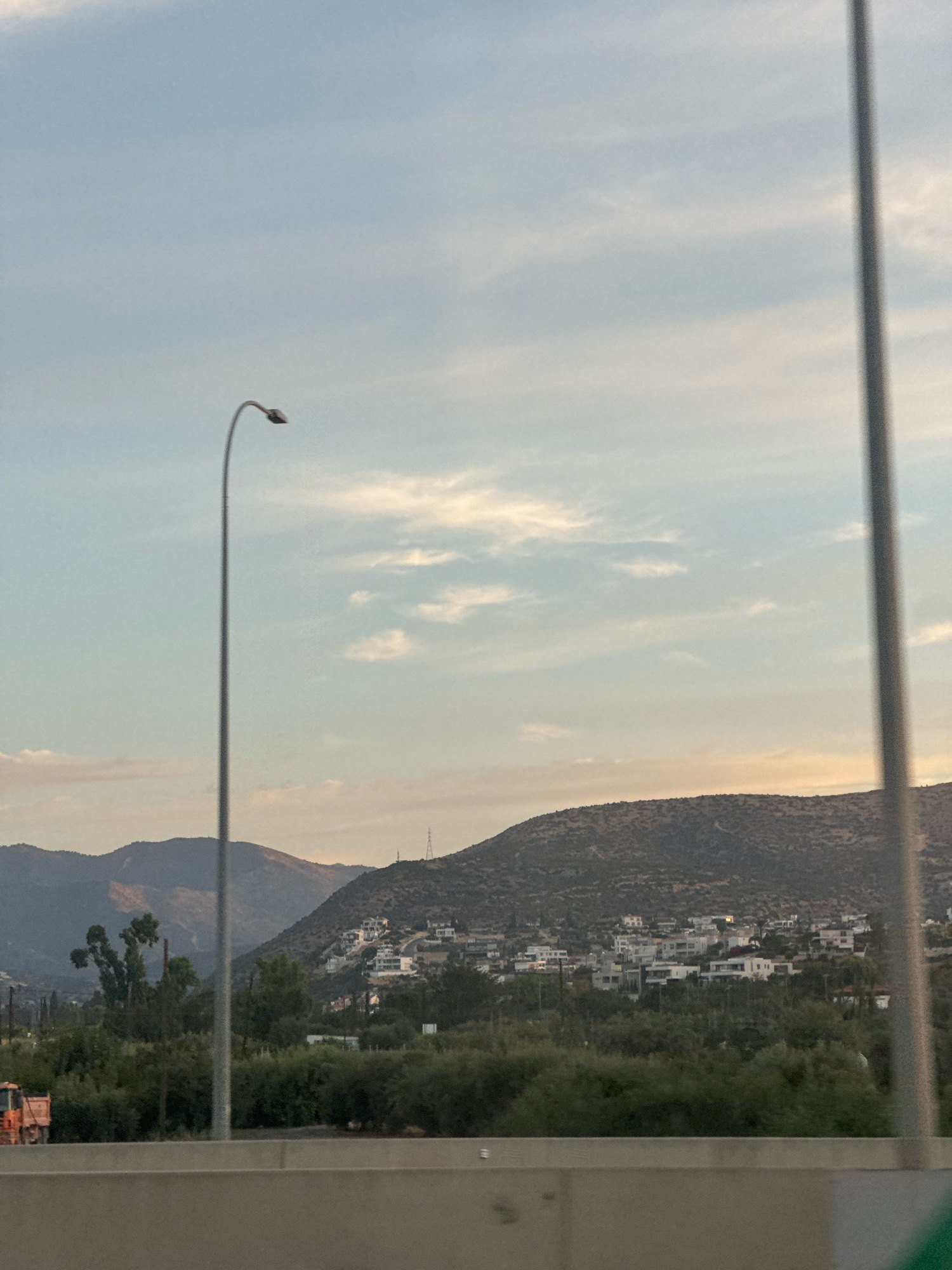 The image captures a view of a hillside settlement in the city of Germasogeia Municipality, Cyprus. The settlement, characterized by its white buildings, is nestled on a sloping terrain covered with vegetation. In the foreground, a concrete barrier runs horizontally across the image, suggesting the perspective is from a road or elevated viewpoint. A tall streetlamp with a curved arm stands to the left of the frame, its pole extending upwards into the hazy sky. The sky itself is a pale blue, with wisps of clouds and a hint of sunset colors near the horizon. In the distance, the hills are covered in dry, scrubby vegetation. To the far left, a section of an orange construction vehicle is visible. The overall scene depicts a semi-rural or suburban landscape under a twilight sky.