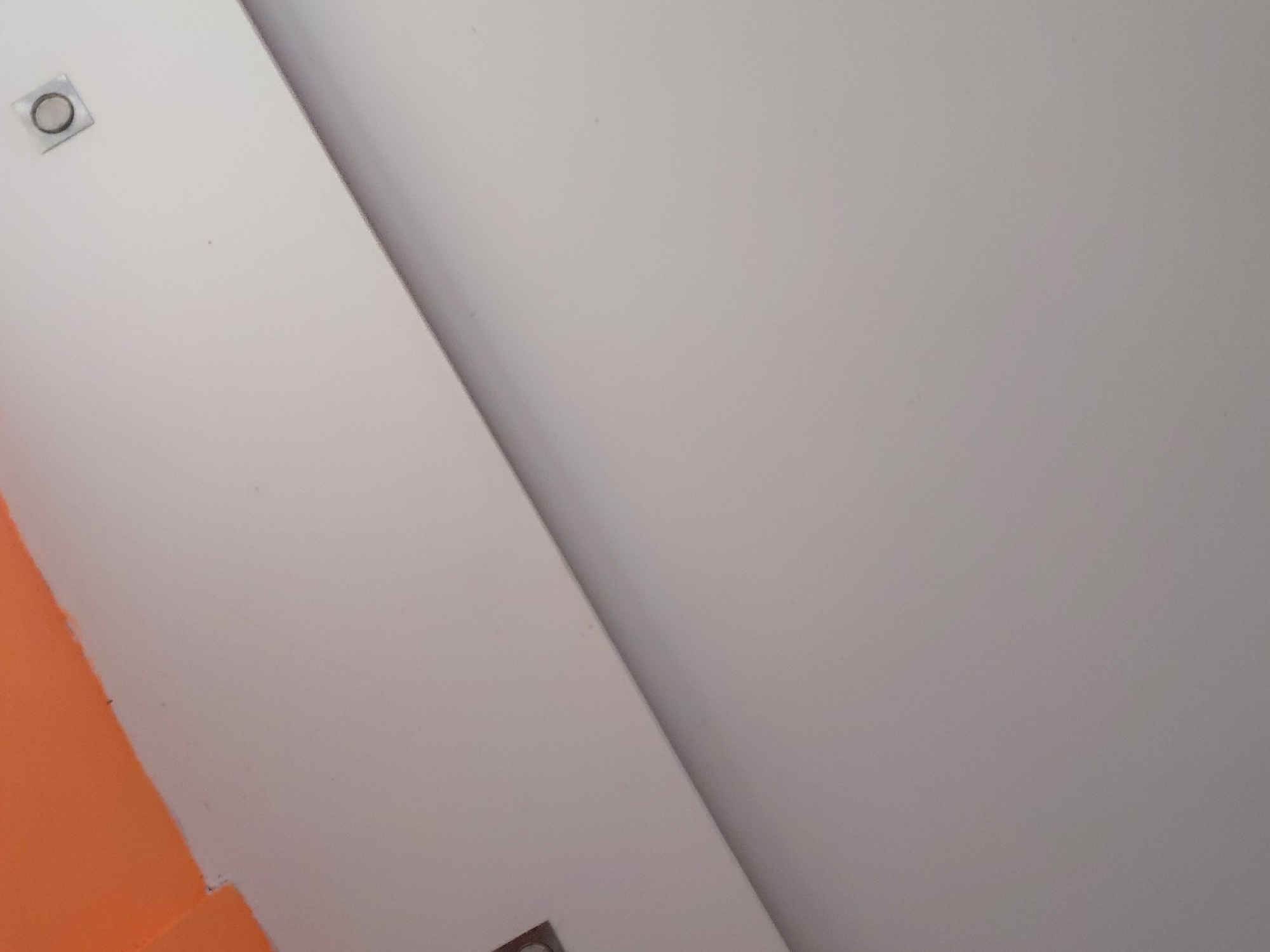 The image presents an abstract composition dominated by architectural surfaces in shades of white, light grey, and orange. The scene is primarily divided by a prominent diagonal line running from the top left to the bottom right, separating a brighter white area on the left from a slightly darker, lighter grey area on the right. This diagonal line suggests a corner or a significant change in surface plane, possibly a wall meeting a ceiling or another wall.
In the upper left quadrant of the brighter white surface, a small, circular metallic object is visible. It appears to be a recessed fixture, possibly a light or a sensor, with a dark ring framing a lighter, reflective center.
Towards the bottom left corner, an irregular section of a vibrant orange surface is visible, sharply contrasting with the adjacent white surface. A dark, thin line marks the boundary between the white and orange areas, suggesting a painted edge or a seam between materials.
The lighting is soft and even, without harsh shadows, making it difficult to determine if it's natural daylight or artificial illumination. There are no people, activities, or discernible text present. The overall impression is one of geometric simplicity and a focus on color and form. No specific location cues are visible within the image to confirm the provided context of Santa Ana, Venezuela.