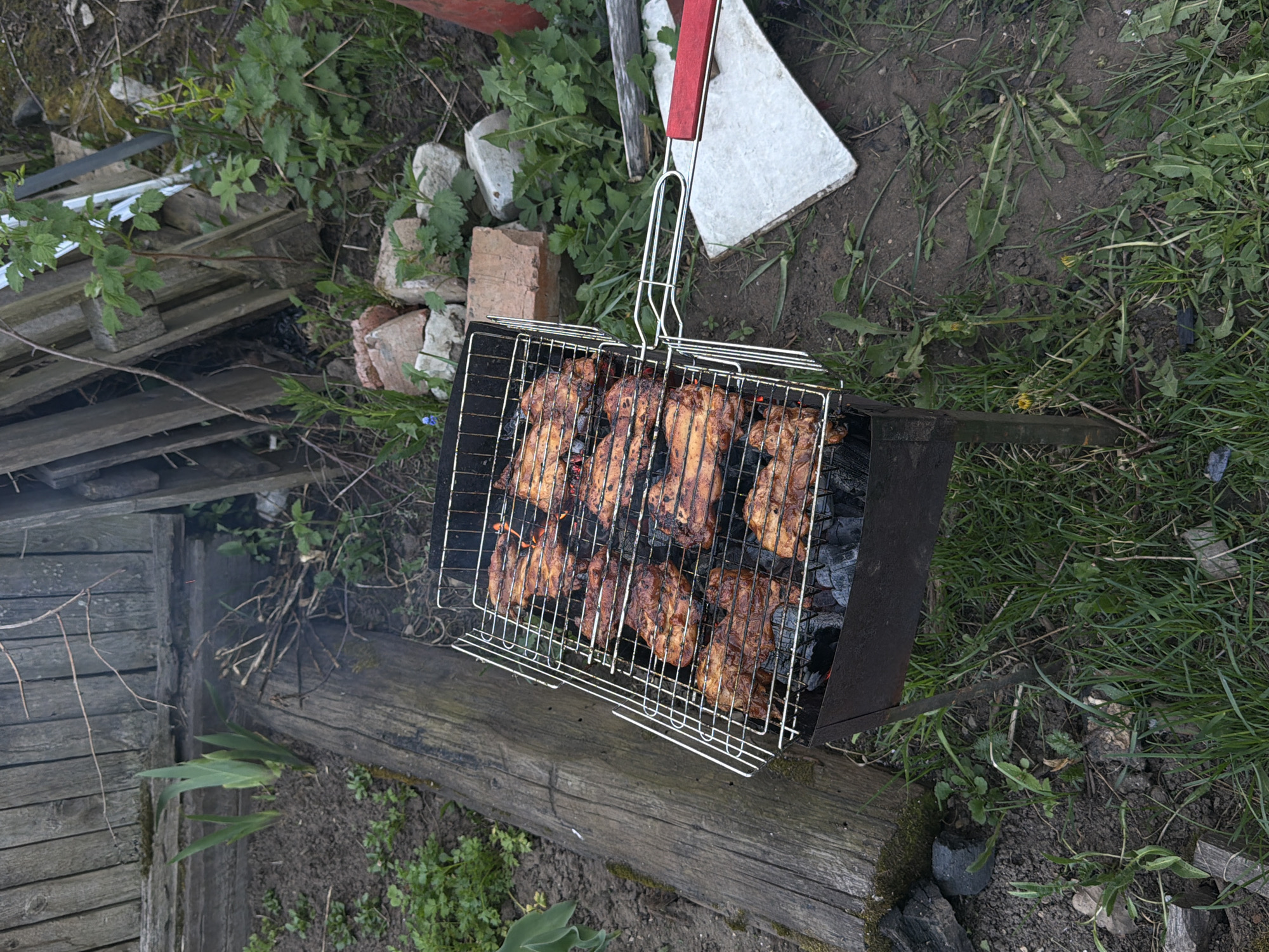 The scene depicts a grill with meat cooking over coals in an outdoor setting. The grill is rectangular, metallic, and appears to be resting on a piece of wood. The meat, which appears to be chicken, is visibly browned and cooking in the grill basket. Surrounding the grill are various elements, including grass, leaves, dirt, pieces of wood, bricks and a white tile. The location of the scene is Perm, Russia. No people are visible in the image.