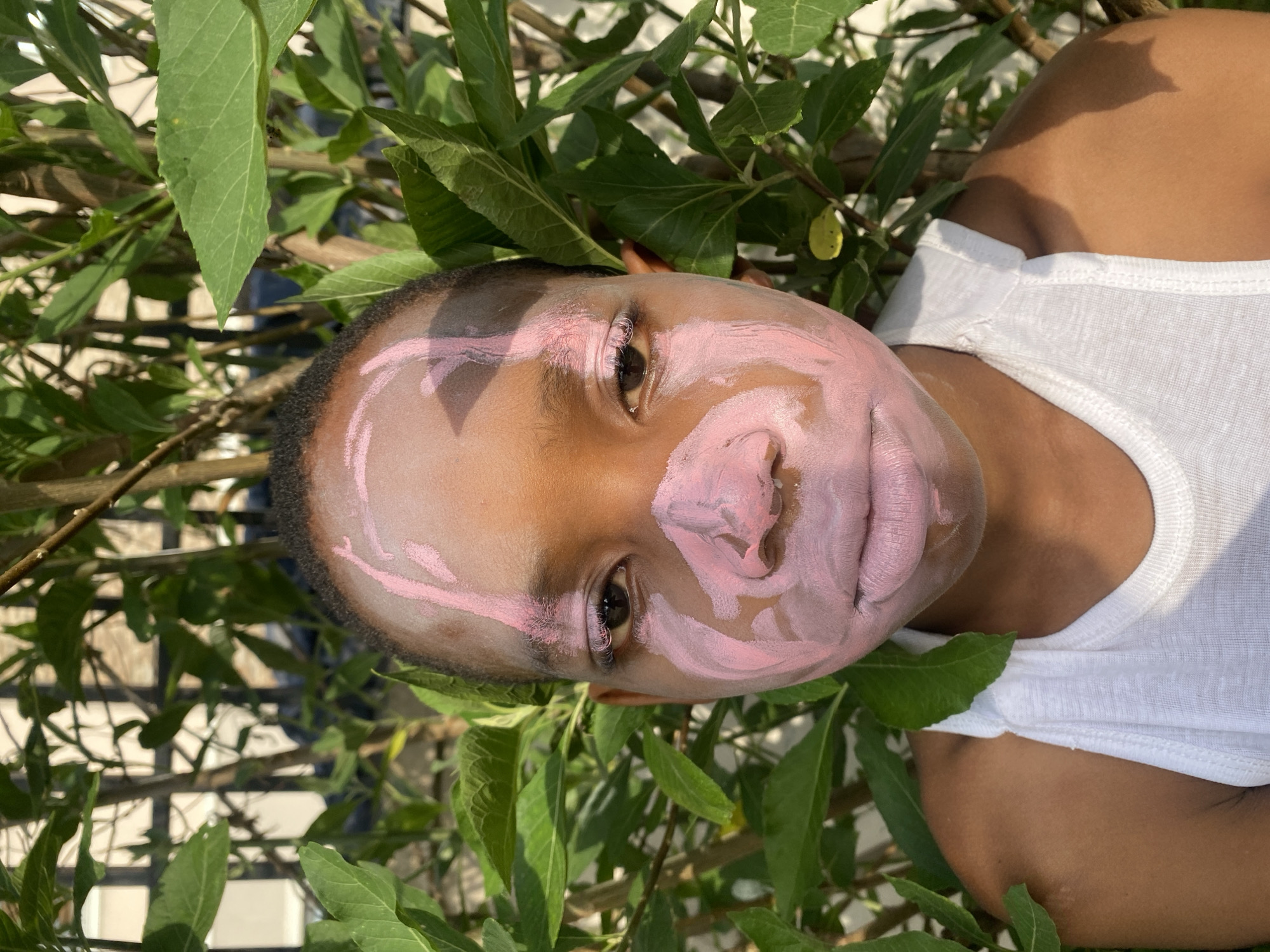 A young individual with dark skin is lying on their back amidst green foliage. The person's face is partially covered in pink paint. They are wearing a white tank top. In the background, a fence and leaves are visible. The location is Iranla, Nigeria.
