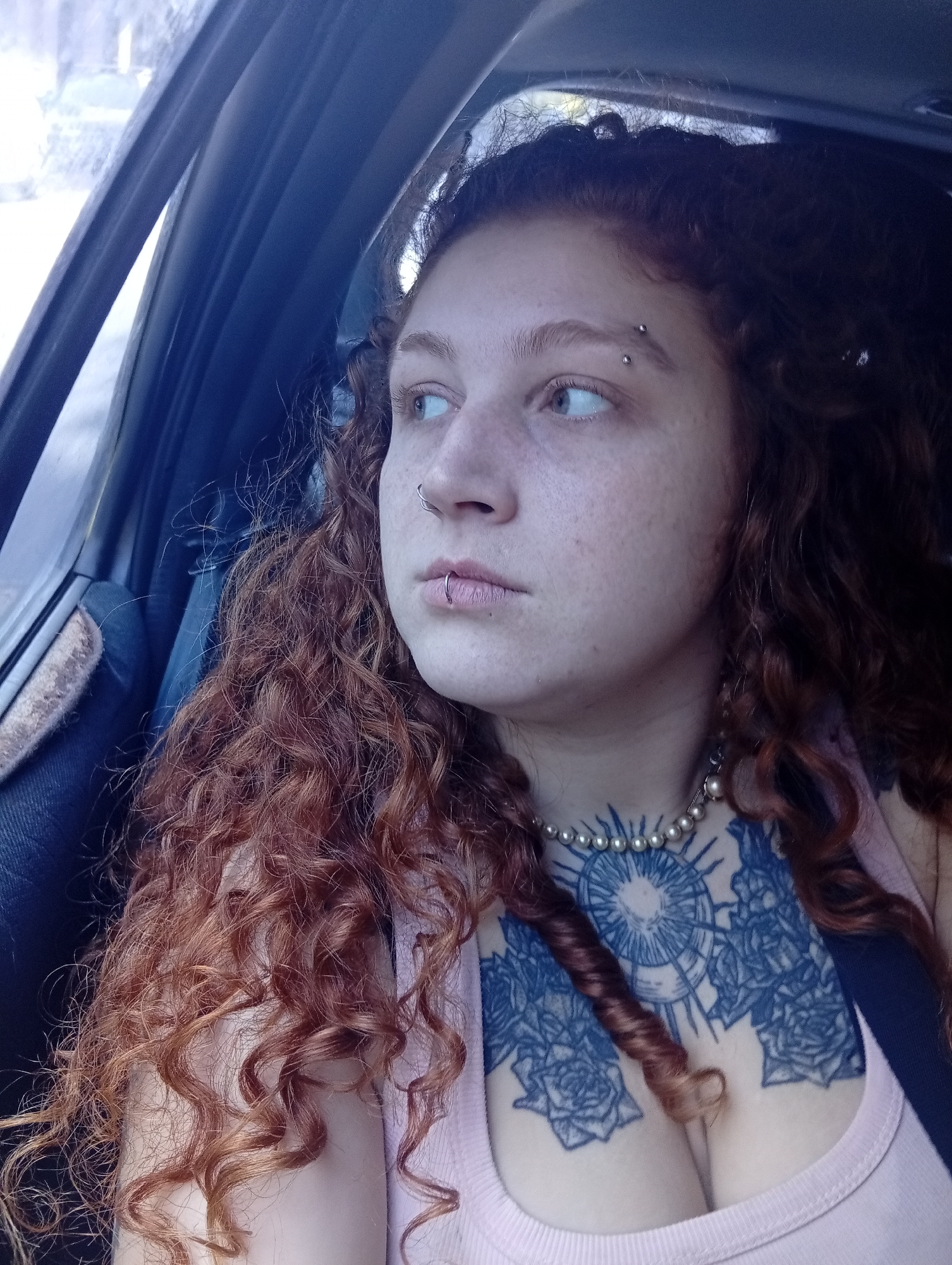 A medium shot depicts a woman seated inside a car, gazing out of the window. She has light skin, freckles, and auburn curly hair. Piercings adorn her left eyebrow, nose, and lower lip. A pearl necklace encircles her neck, positioned above a visible chest tattoo with floral and sunburst motifs. She wears a light pink tank top. The car's interior, featuring dark blue upholstery and trim, is visible. The scene is captured during daylight. The location of the city of Buenos Aires, Argentina, cannot be determined from the image.