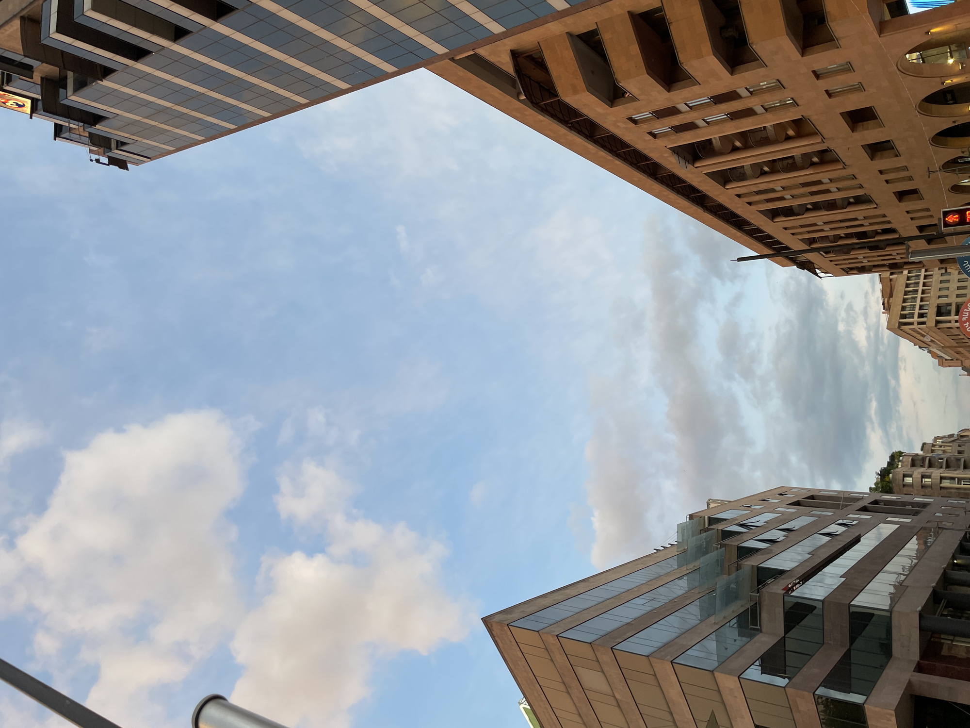 The image shows a low-angle view of buildings and sky in Yerevan, Armenia. At the top left is a section of a modern building with a grid of windows. To the right is a section of a building with an elaborate facade including archways and recessed windows, and a traffic light. In the foreground, at the bottom right, is a section of another modern building with angled sections and large windows. The sky is pale blue with scattered clouds.