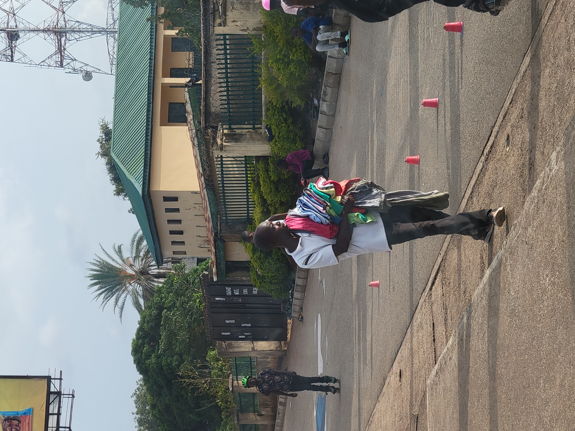 A man in a white t-shirt walks along a street carrying a bundle of clothes over his shoulder, moving past a series of small, orange cones. Another person with a green helmet and wearing dark clothing is standing on the street. In the background, there's a low building with a green roof, surrounded by a fence and some greenery. The scene is set in Jos, Nigeria, as suggested by the architectural styles and vegetation present. A communication tower looms overhead.
