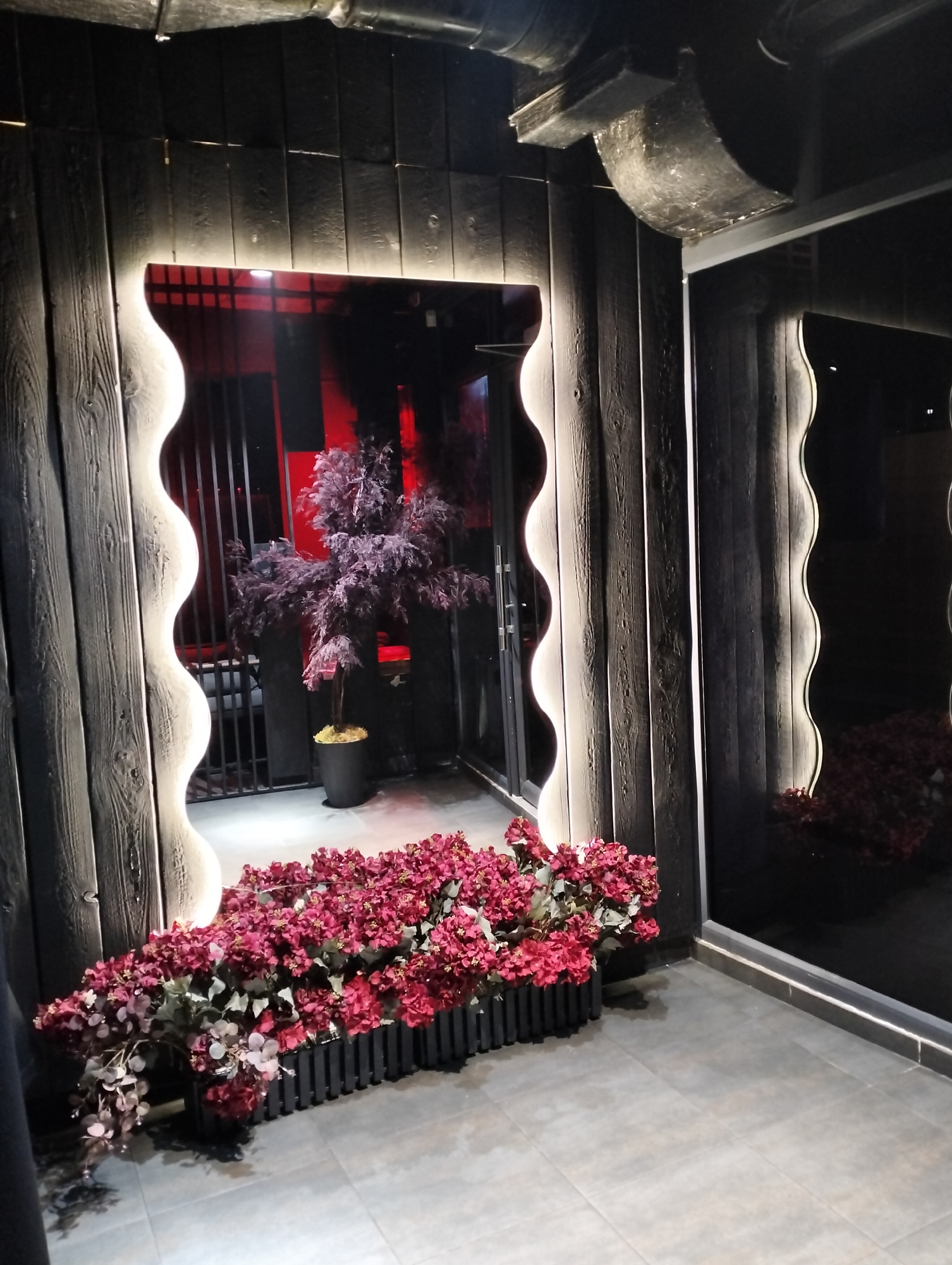 A dimly lit interior or exterior space features dark wooden walls with vertical planks. Two rectangular openings, possibly windows or doorways, are bordered by wavy, illuminated frames. Red flowers are arranged in black planters at the base of the left opening. Inside the left opening, a small tree stands in a black pot. The space reflected in the right opening shows similar floral arrangements. The floor is tiled with large, gray tiles. The ceiling is partially visible, revealing dark elements, possibly pipes or structural components. The location is the city of Ivano-Frankivsk, Ukraine.