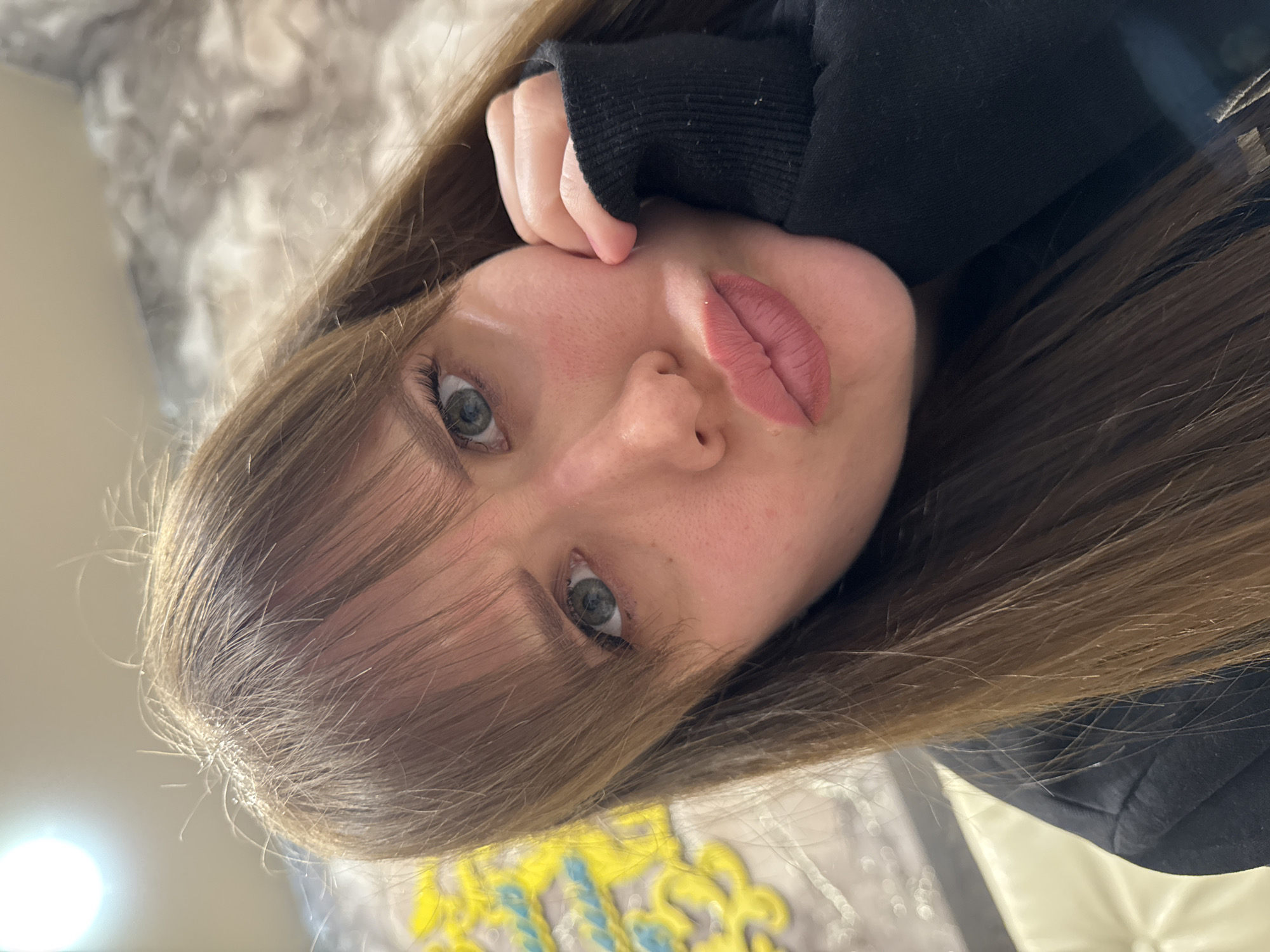 A close-up, angled portrait features a young woman with long, straight, medium-brown hair with lighter strands, partially covering her forehead with bangs. She has striking blue-green eyes that look directly at the viewer. Her lips are full and appear to have a soft pink or nude lip product. Her fair skin shows natural texture. She is wearing a black long-sleeved garment, likely a hoodie or sweatshirt, with the cuff of the sleeve pulled up and resting near her temple. Her hand, with fingers slightly curled, also rests against her face.
The woman's expression is neutral and calm, possibly contemplative. Her posture suggests a relaxed state, perhaps resting. The background is softly blurred, indicating an indoor setting, possibly a bedroom or living room, with indistinct patterns and shapes, including hints of yellow and blue, suggesting a blanket, pillow, or upholstered furniture. A soft light source creates bokeh effects in the upper left background. No specific time of day or weather is discernible. There is no visible text in the image. While the location context is Chaikovsky, Russia, no visual cues within the image itself confirm this specific geographical location.