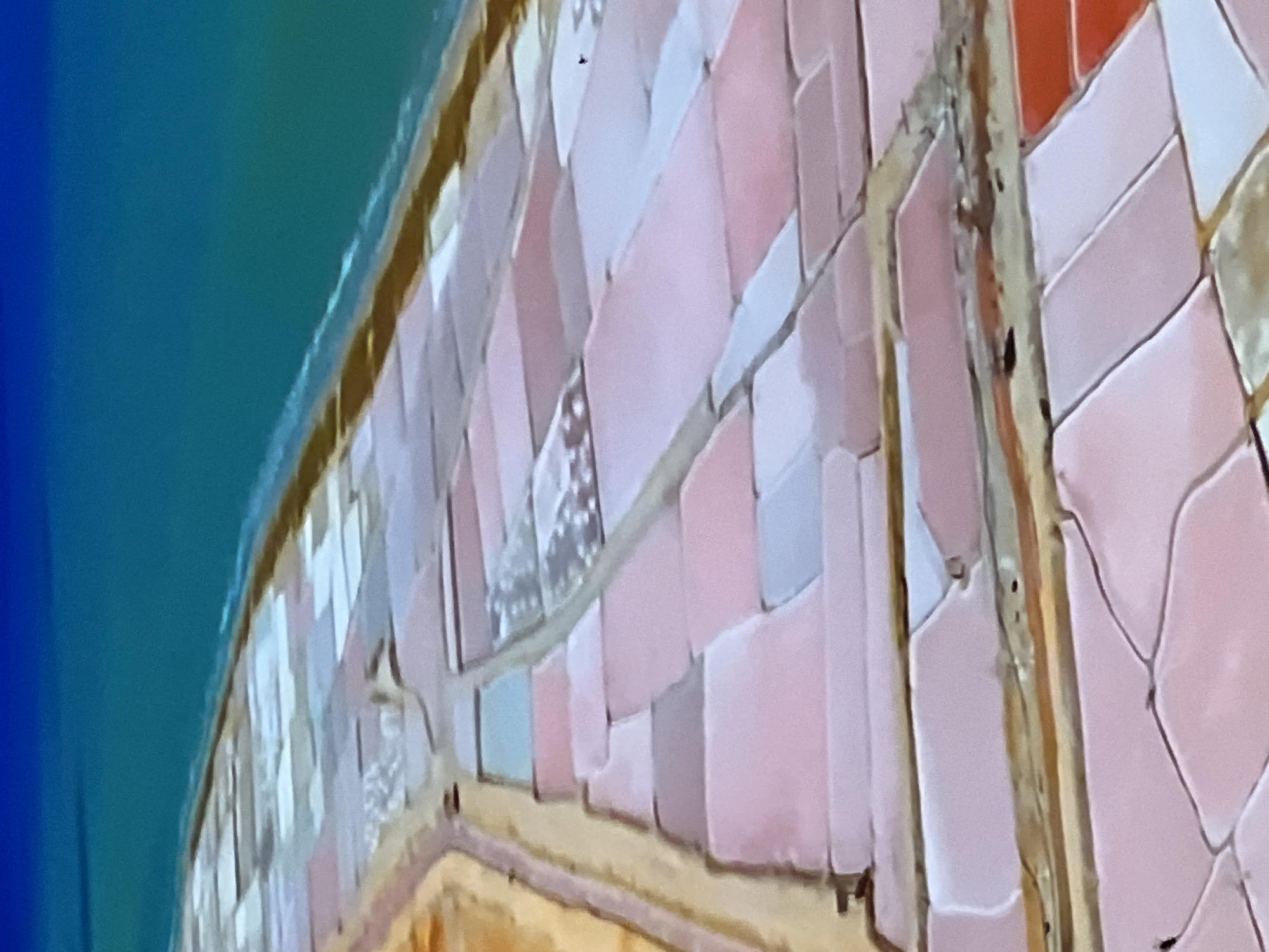 The aerial image displays a salt evaporation pond system. The ponds are rectangular and arranged in a grid-like pattern, separated by earthen dikes. The water within the ponds exhibits varying colors, ranging from pale pink and white to shades of blue and orange, indicating different stages of salt concentration and crystallization. In the upper portion of the image, a body of turquoise water, likely the ocean, is visible, bordering the salt ponds. A strip of sandy terrain is present between the ponds and the ocean. The scene depicts an industrial or agricultural process related to salt production. There are no people or specific landmarks visible that would definitively place the location in Casablanca, Morocco, or any other specific city.