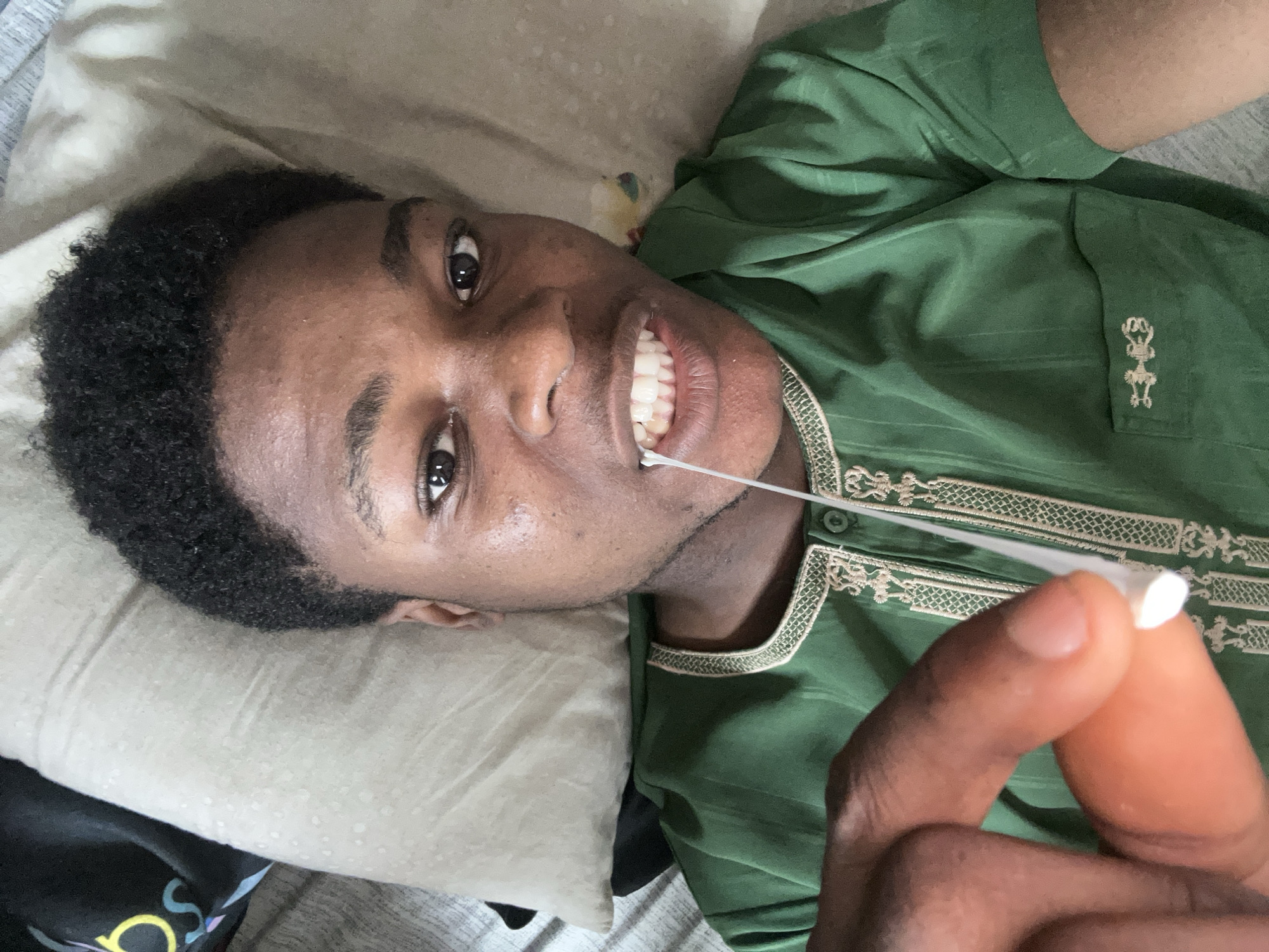 The media file shows a close-up, top-down view of a young Black person lying on a bed, holding a Q-tip near their mouth. The person is wearing a green, embroidered shirt and has dark, curly hair and dark eyes. Their mouth is slightly open, revealing their teeth, and the Q-tip appears to be touching their teeth. The background consists of a light-colored pillow and bedding. The lighting suggests it could be daytime indoors. The person's expression is neutral or slightly amused. There is no visible text in the image. The scene appears to be in a bedroom setting in Ihiagwa, Nigeria.