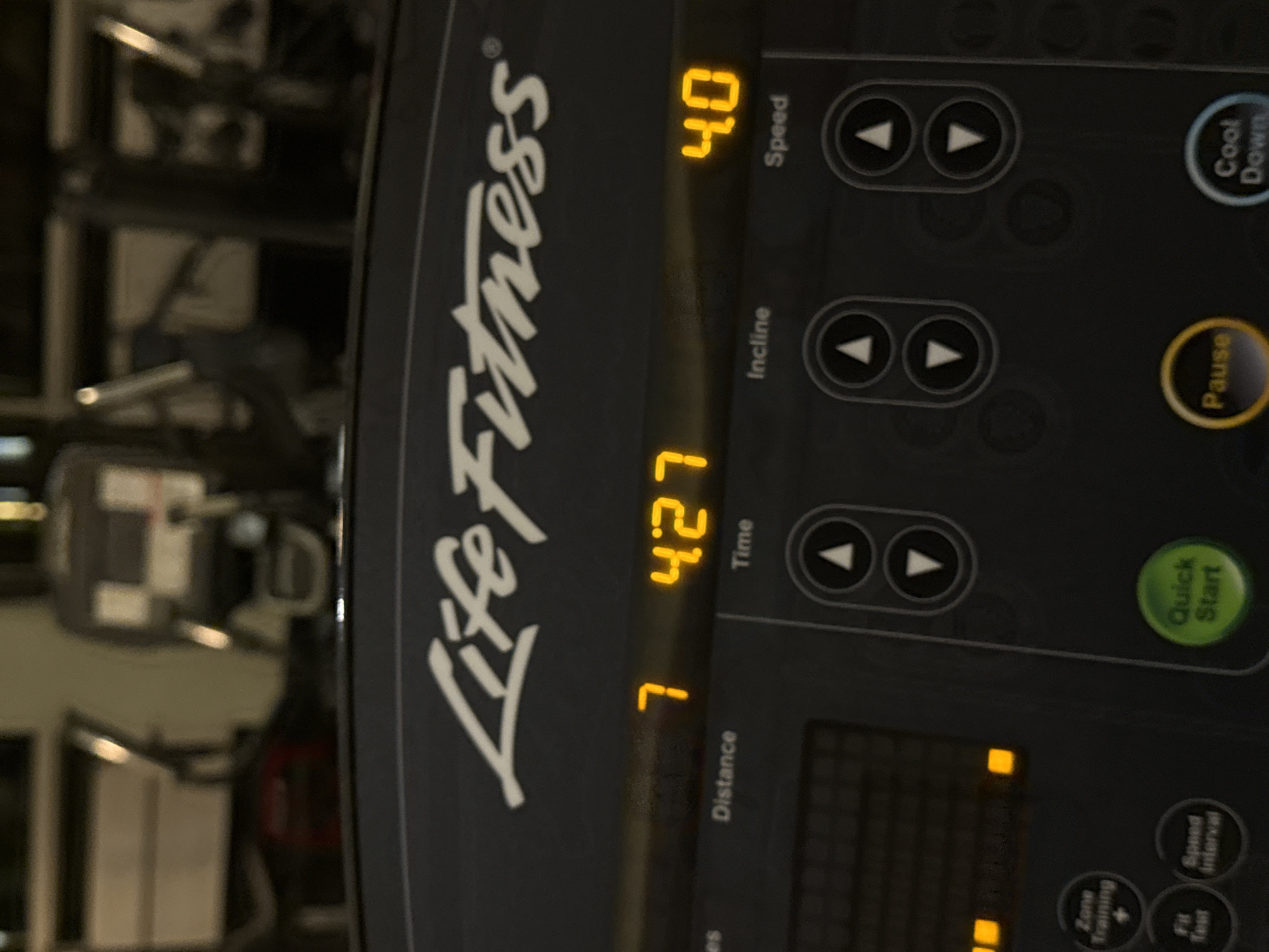 The image displays a close-up view of the digital console of a Life Fitness treadmill, oriented with a 90-degree counter-clockwise rotation. The console, dark gray in color, features the prominent "Life Fitness" logo in white script. Bright orange-yellow digits on the display indicate various workout metrics. The "Speed" is shown as "4.0", and the "Time" elapsed is "4:27". Below a partially visible label for "Distance", the number "7" is displayed.
The console is equipped with several control buttons. On the right side, circular buttons are labeled "Cool Down", "Pause", and "Quick Start". Further down, smaller circular buttons are visible for "Zone Training +", "Fit Test", and "Speed Interval". Sets of arrow buttons are present for adjusting "Speed" and "Incline". A rectangular grid-like display, with several segments illuminated, likely indicates program progress or intensity. The background is blurred, revealing indistinct shapes of other fitness equipment, suggesting the setting is a gym or fitness center in Mexico City, Mexico. No people are visible, and no specific time of day or weather conditions can be determined from the image.