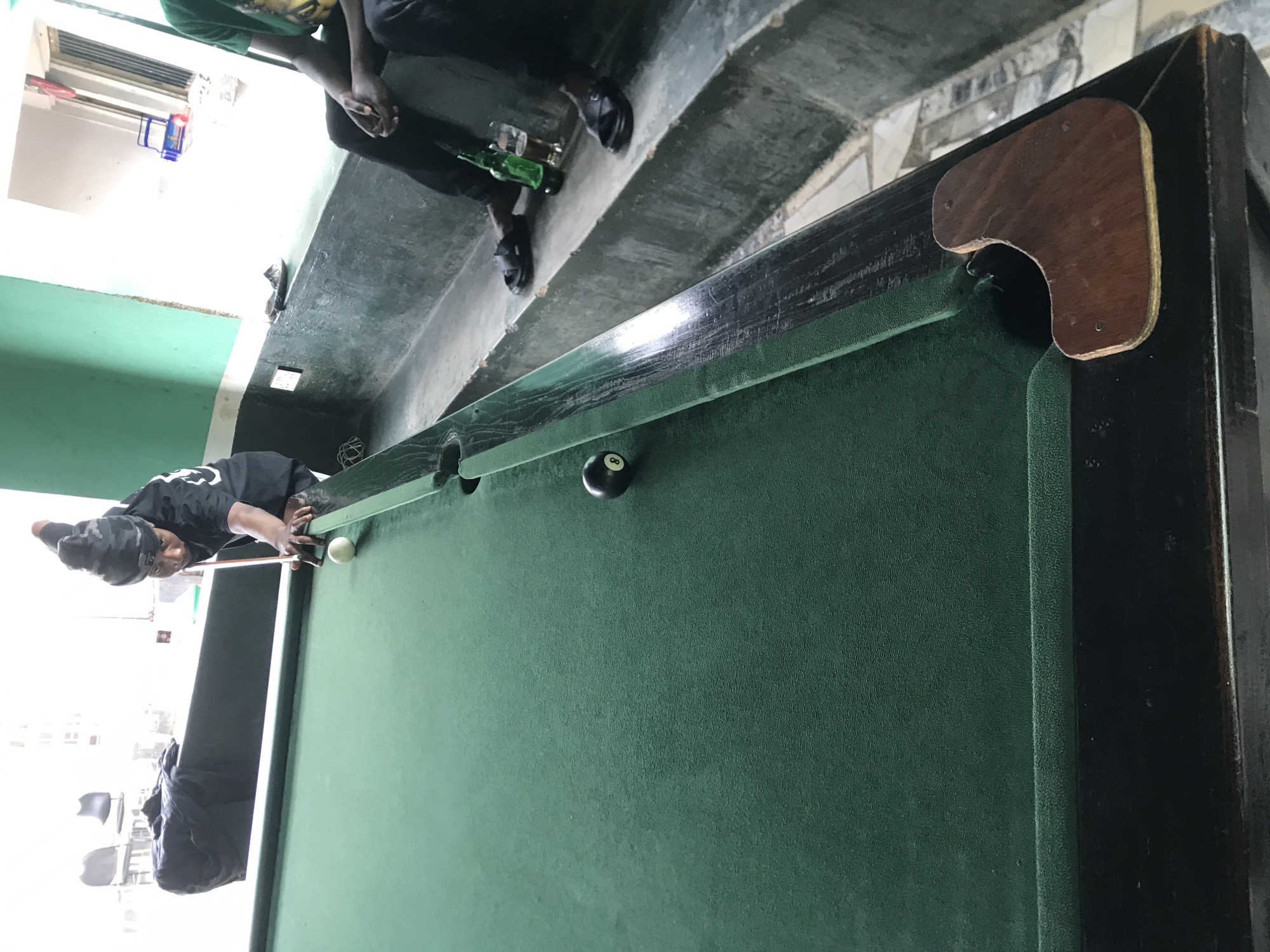 The image captures a casual scene at what appears to be a pool hall or recreation area, potentially within or near the New Stadium in Nigeria. The primary subject is a person, wearing a black beanie or headwrap and a black long-sleeved shirt with white graphics, leaning over a green-felt pool table to take a shot. They are holding a pool cue, aligning it with the cue ball, which is positioned to strike another ball (possibly an 8-ball, which is visible in a pocket further down the table).
Another person is partially visible in the background, standing on concrete steps or a raised platform behind the pool table. This individual wears a green t-shirt and dark trousers, and their feet are adorned with sandals. A green glass bottle, possibly of a beverage, and a drinking glass are placed on the steps near this person.
The setting features rough, dark concrete flooring and steps leading upwards, suggesting an outdoor or semi-outdoor covered space. The background is brightly lit, indicating it is daytime, with glimpses of a white wall and a small, brightly colored children's basketball hoop visible. The pool table itself is dark-framed with wooden corner accents. No discernible text is clearly visible on any objects or clothing. The atmosphere seems relaxed and focused on the game.