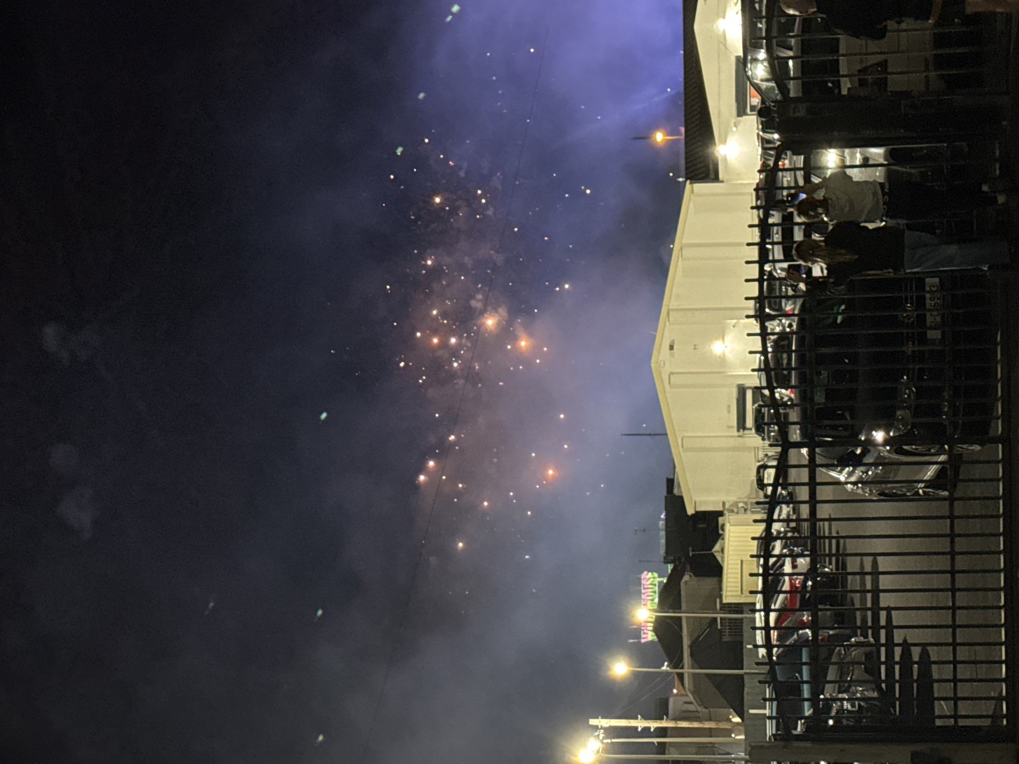 The image depicts a nighttime scene in Tramore, Ireland, likely during a fireworks display. In the foreground, a black metal gate separates the viewer from a parking lot filled with various cars. Behind the gate, several vehicles are visible, with some parked in rows. Two individuals, a woman with long blonde hair and another person with lighter hair, stand behind the gate, facing away from the camera and towards the sky. Both appear to be holding up their phones, possibly to record the event. In the middle ground, several buildings are present, including a white structure with a flat roof and a building with a neon sign that reads "Wild House." The sky is dark and filled with smoke and numerous bright, colorful fireworks. The fireworks are scattered across the upper portion of the image, creating a vibrant spectacle against the dark sky. Streetlights are visible in the distance, illuminating parts of the surrounding area.