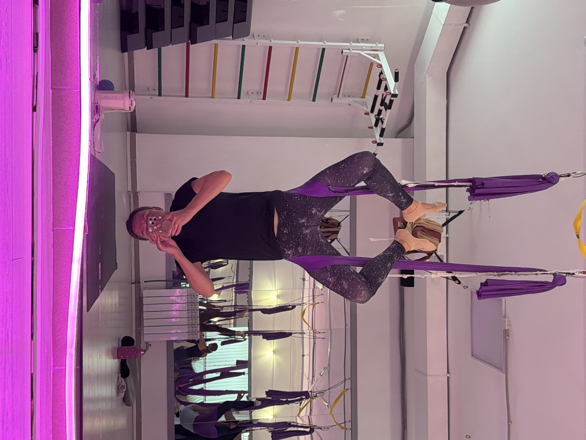 A woman is performing aerial yoga in a studio. She is suspended upside down from a purple aerial hammock attached to the ceiling. Her legs are wrapped in the fabric, and her arms are reaching up, holding onto the hammock. She is wearing patterned leggings and a dark t-shirt. In the background, there are mirrors, other aerial hammocks, and exercise equipment. The studio is illuminated with bright pink lighting from the floor. A Swedish ladder is visible on the wall to the left, and a yoga mat with a water bottle and other items is on the floor in the foreground. The presence of aerial yoga equipment and a Swedish ladder suggests this is a fitness or dance studio. The location is identified as Chaikovsky, Russia.