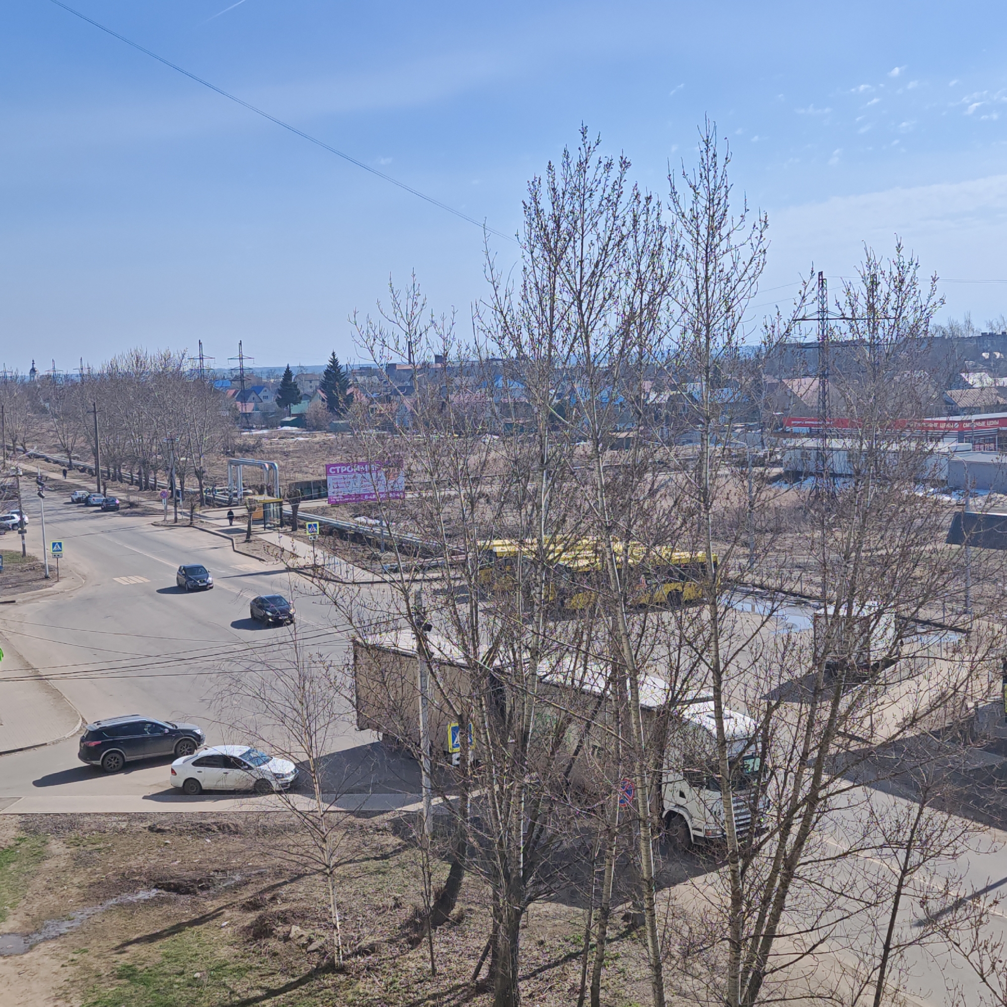 The image depicts a street scene in Rostov, Russia, from an elevated perspective. In the foreground, bare trees frame the view of a street intersection. A black SUV and a white car are positioned at the corner. On the road, a dark sedan and a silver sedan are driving. Across the street, a billboard and a bus stop are visible. A yellow bus is parked nearby. The background shows residential buildings and industrial structures under a clear blue sky with power lines.