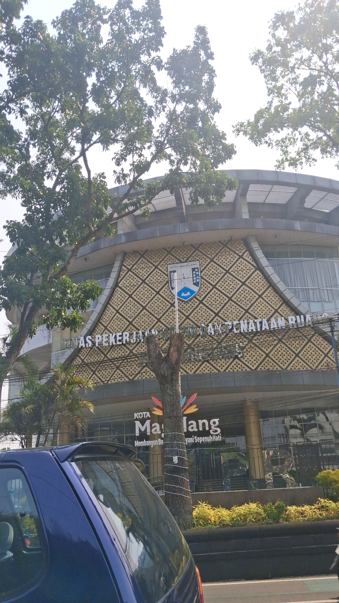 The image shows the facade of a building in Magelang, Indonesia. The building has a decorative pattern on its upper section and a large sign that reads "KOTA MAGELANG" with smaller text below. A blue car is visible in the foreground. Trees and foliage surround the building. The sky is bright, indicating daylight. The building appears to be an office or public building.