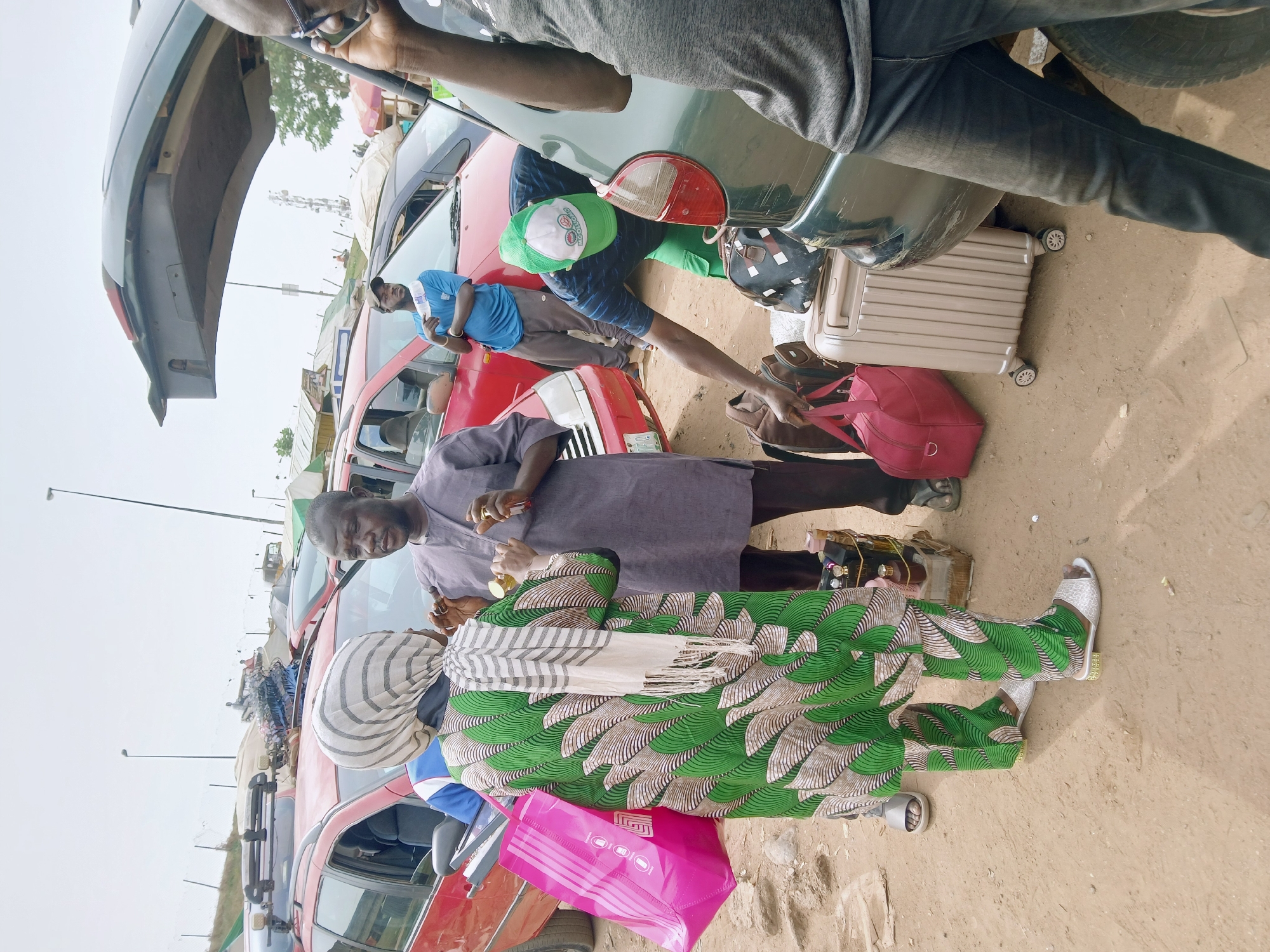 The image shows a street scene in an unknown city with several parked cars and people. A person in a green cap and shorts is reaching towards a pink bag held by a person in a gray shirt. The person in the green cap is standing near a suitcase and a dark bag. A woman in a green and white patterned outfit stands to the left. Another man stands behind her, wearing a purple shirt. The trunk of a green car is open. Cars are lined up behind them, and other people are visible near the cars. The ground is a light brown color, and the sky is overcast.
