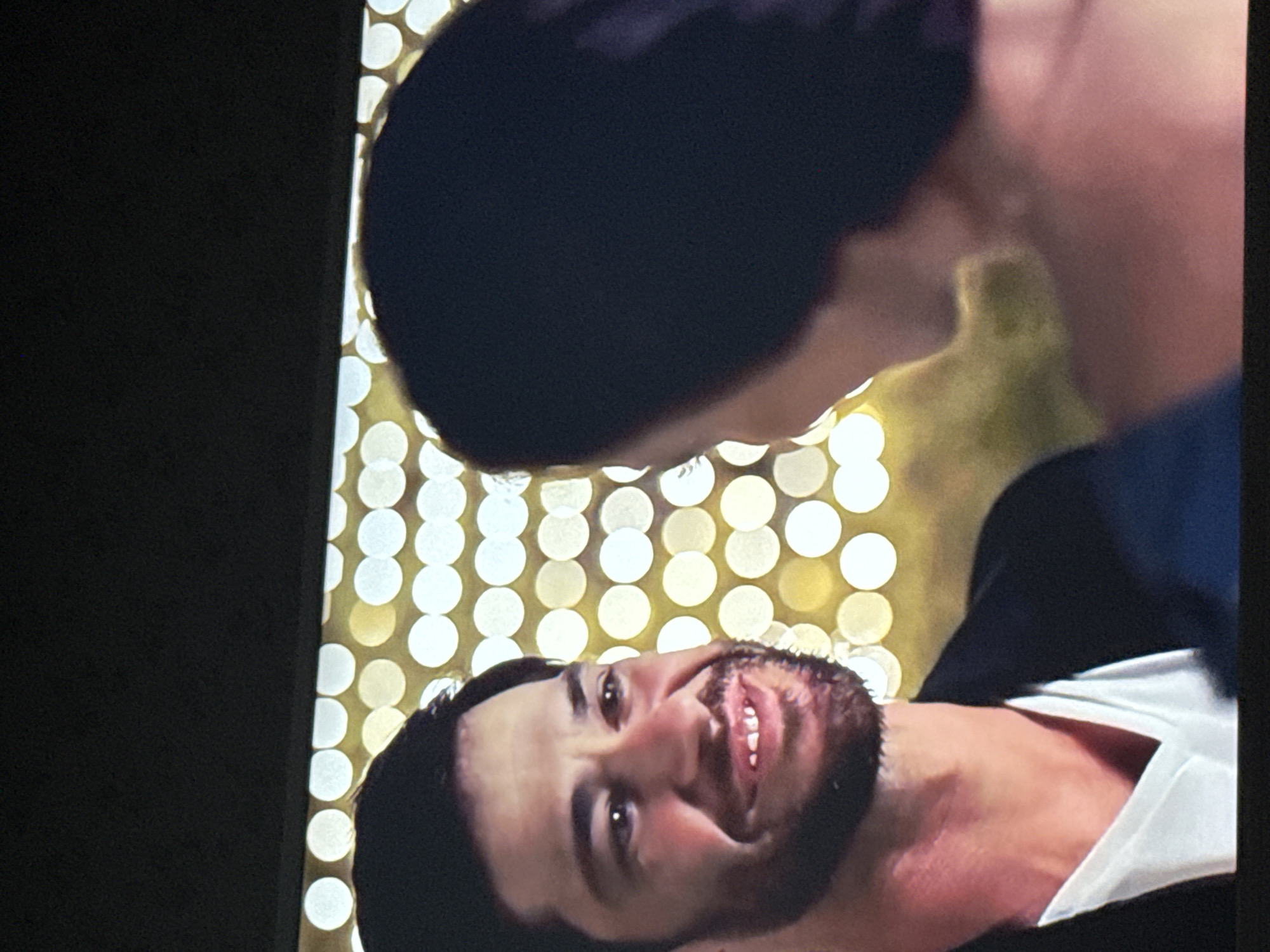 The image depicts two people, one lying down and facing upward, the other leaning over them. The person lying down has dark hair, a beard, and is wearing a white shirt and dark jacket. They are smiling. The person leaning over has short, dark hair and their face is partially visible. The background is a blur of white and gold circular lights. There is no indication that the image was taken in Mytishchi, Russia.