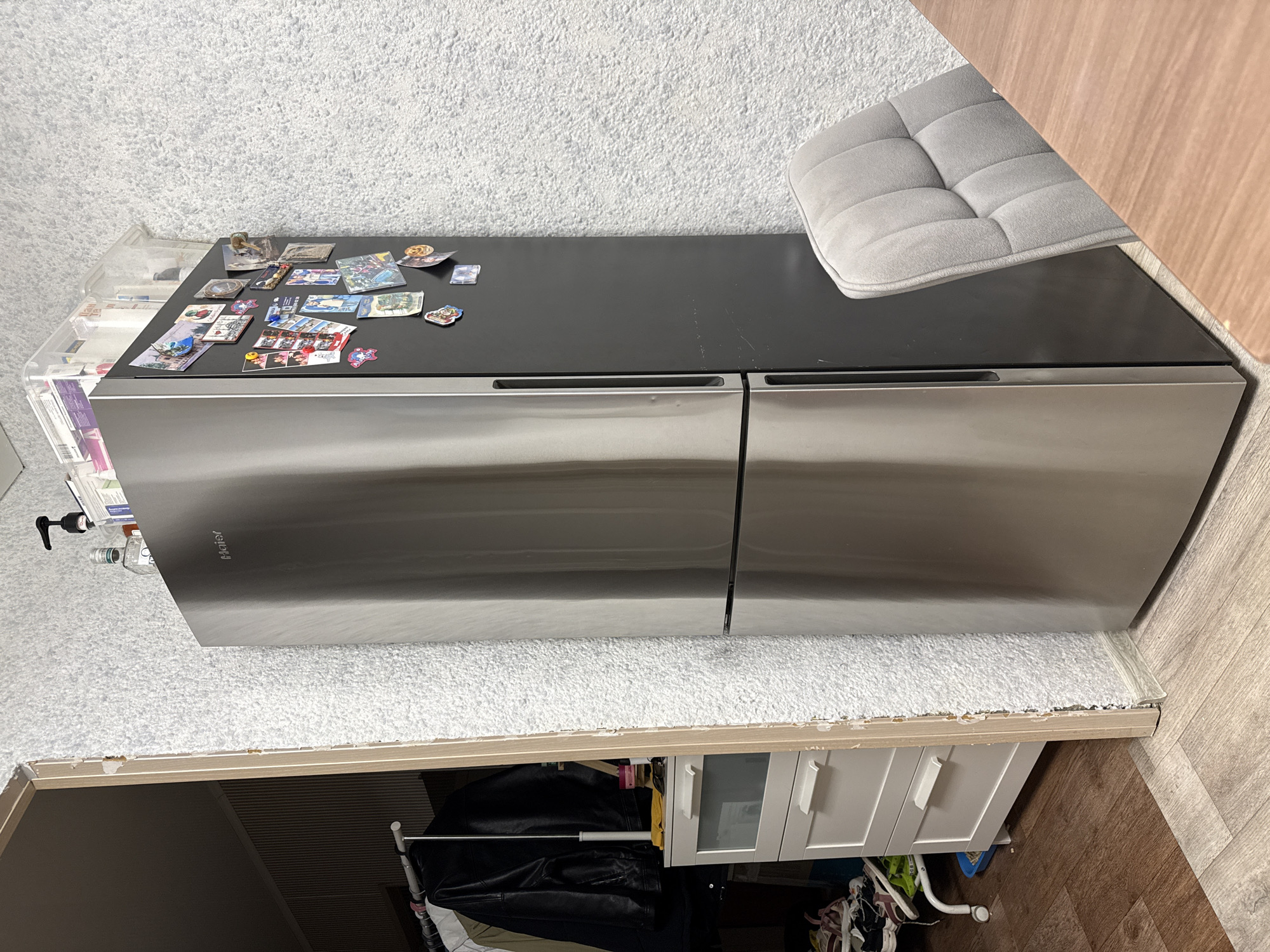 The image shows the top of a stainless steel refrigerator with a black glass top. Various magnets and small pictures are adhered to the black surface. A light gray, tufted cushion is placed on the right side of the refrigerator top, partially obscuring the view of the refrigerator. To the left of the refrigerator, a clear plastic storage bin with multiple drawers is visible, filled with what appear to be papers or cards.
Below the refrigerator, a section of a white cabinet unit with two drawers and two glass-fronted cabinet doors is visible. There are also clothes hanging on a rack in front of the cabinet. The flooring is wood-look laminate.
The scene appears to be indoors, likely in a kitchen or dining area. The overall impression is of a domestic setting, possibly a home. There is no indication of the time of day or weather conditions. The image is taken from a high angle, looking down onto the refrigerator.
Visible text includes "Haier" on the refrigerator door.