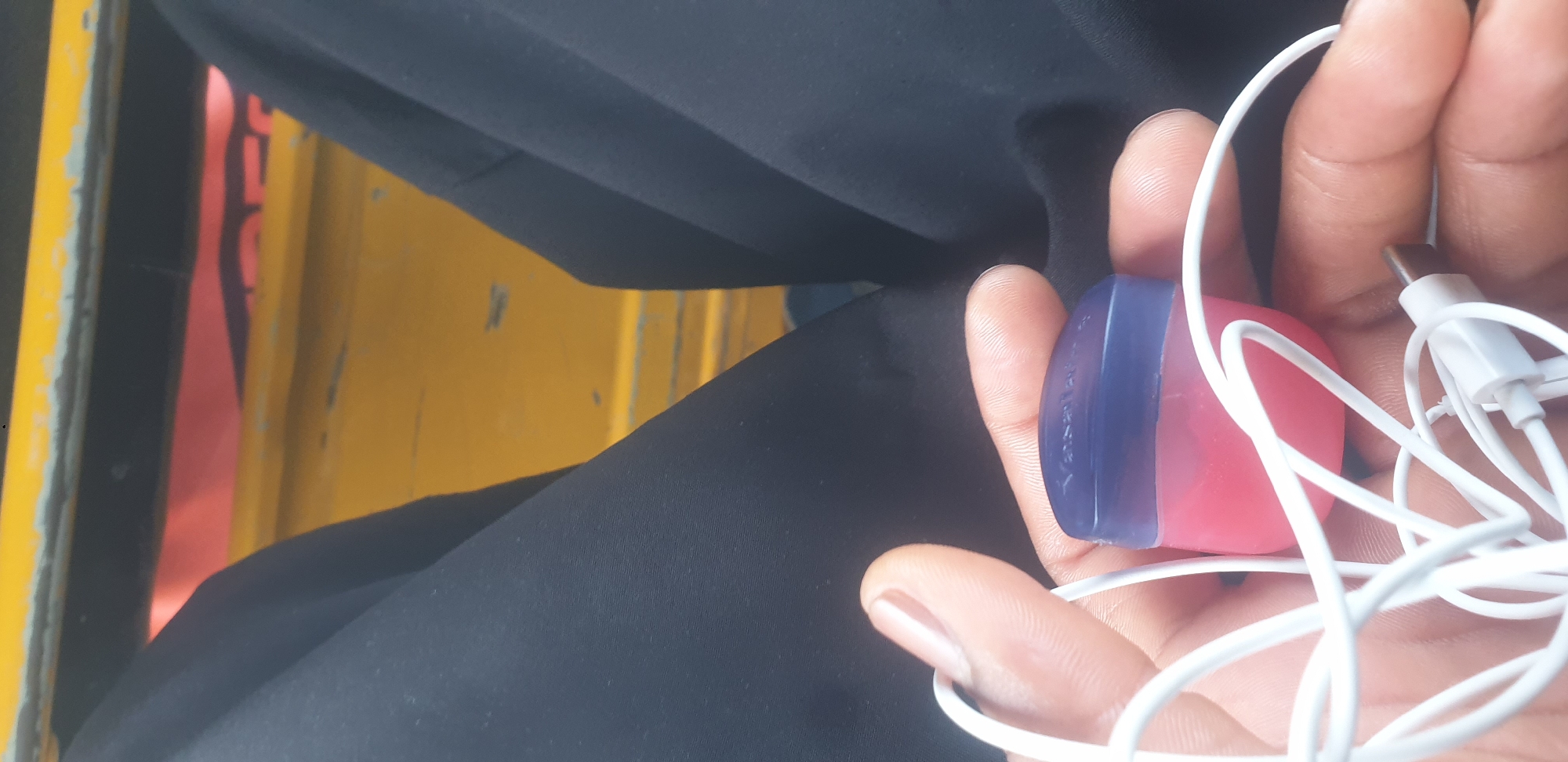 In Barakin Lerawi, Nigeria, a close-up shot focuses on a hand holding a small, round container of Vaseline with a blue lid and pink base. White earbuds are tangled around the container and the fingers. The background is blurred, showing dark fabric and a bright yellow surface, suggesting the person is sitting in a vehicle.
