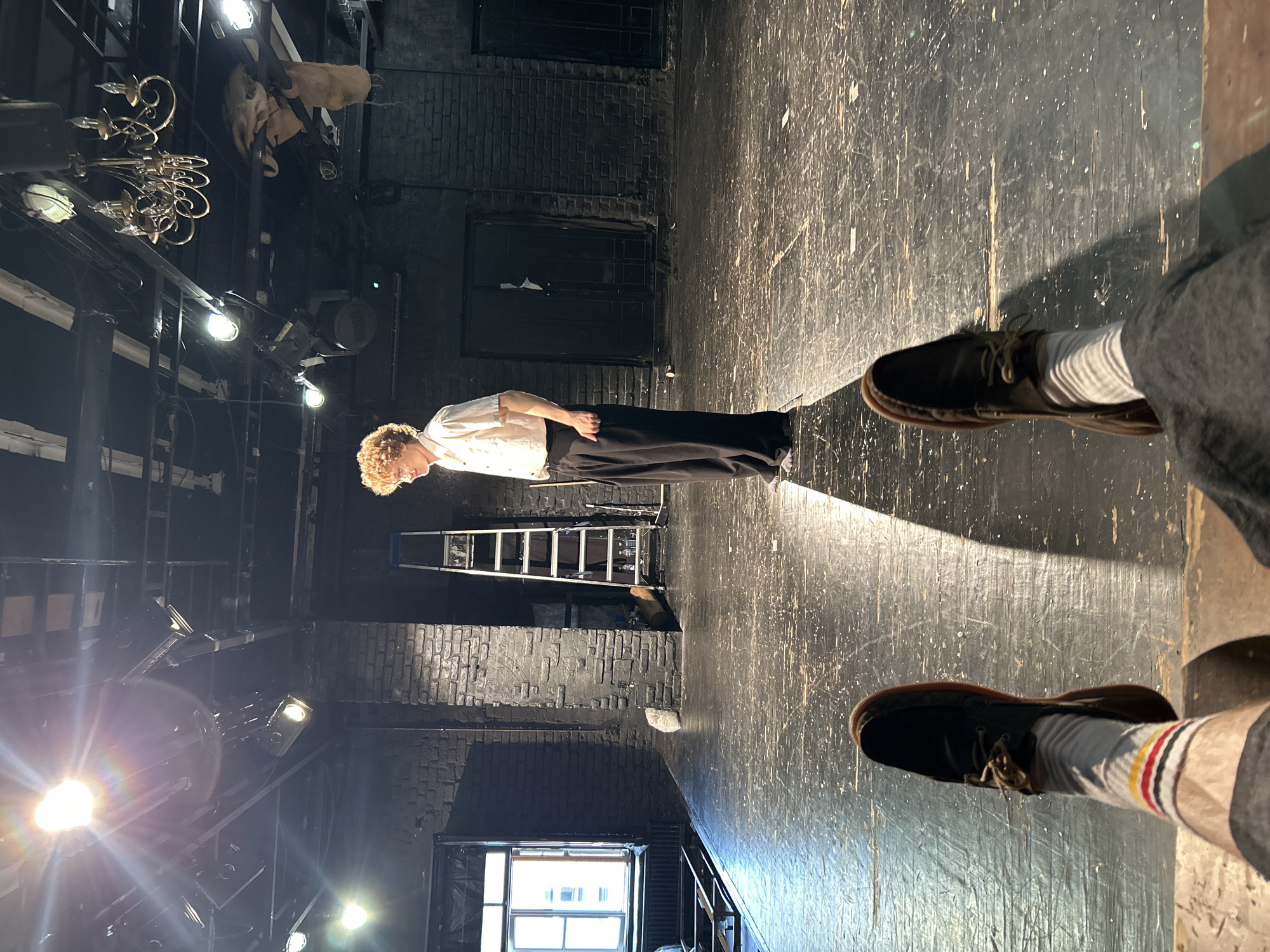 The image shows a person standing on a wooden stage in what appears to be a theater or performance space. The person, with curly blond hair, is wearing a white shirt and dark pants, with their body angled towards the right side of the frame. They are illuminated by a spotlight, casting a shadow behind them.
The stage floor is dark and shows signs of wear, with numerous scuff marks and scratches. In the background, there are brick walls and what looks like a ladder. Above the stage, there are various theatrical lights and rigging, suggesting a performance setup. On the right side of the frame, the feet of another person are visible, wearing dark shoes and striped socks, with their legs crossed.
The overall atmosphere is dimly lit, with the spotlight on the performer being the primary source of light. The scene evokes a sense of anticipation or reflection, possibly during a rehearsal or a performance. There is no visible text in the image. The time of day and weather are not discernible from the image.