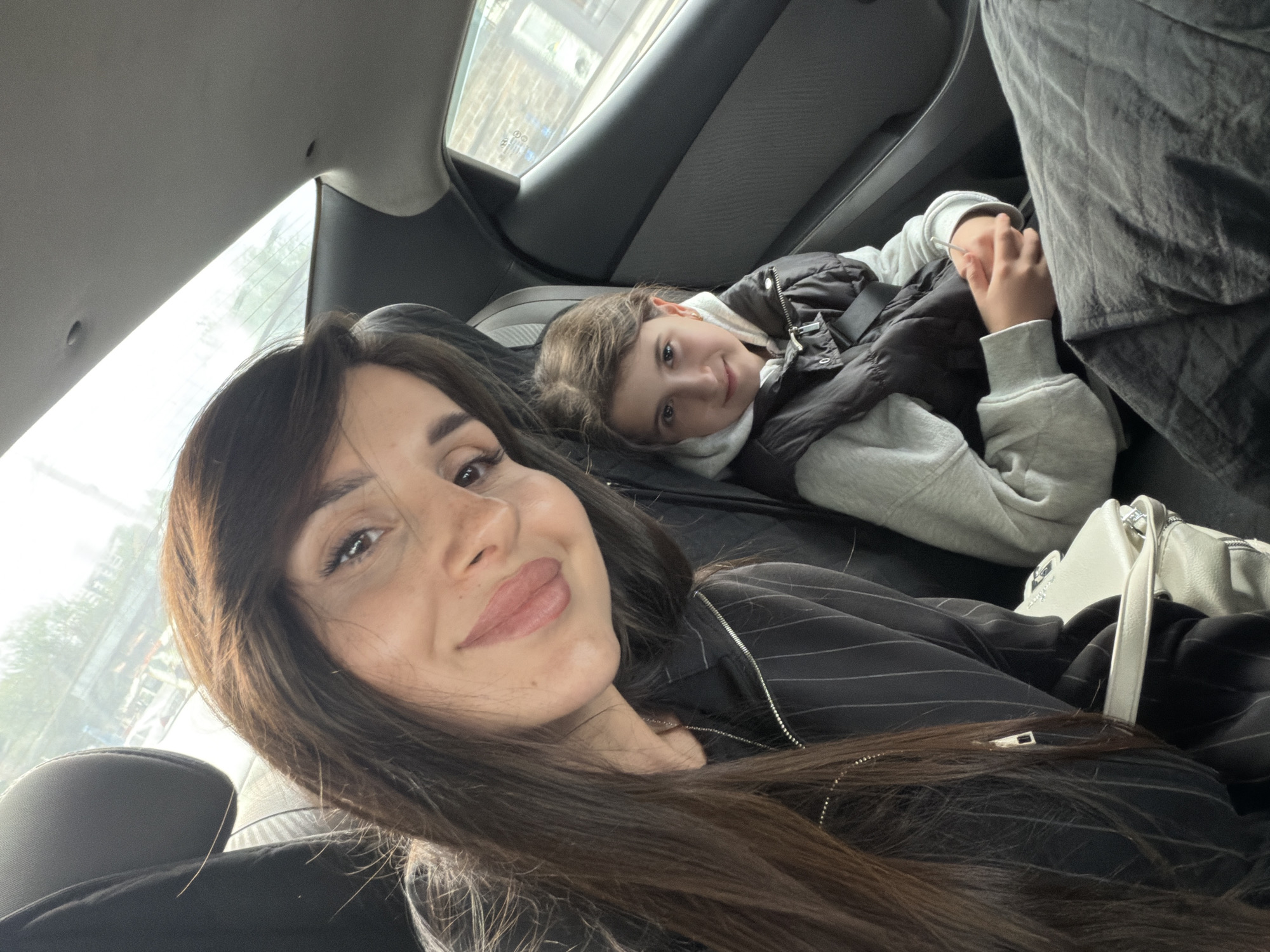 A selfie taken inside a car shows a woman in the foreground with a young girl lying behind her. The woman has long brown hair, brown eyes, and fair skin. She is wearing a black pinstripe jacket. Behind her, the girl is lying down, wearing a grey hoodie and a black vest. A white purse rests near the woman. Through the car window, buildings are visible, suggesting a city environment. Based on the surroundings, the location is likely Yerevan, Armenia.