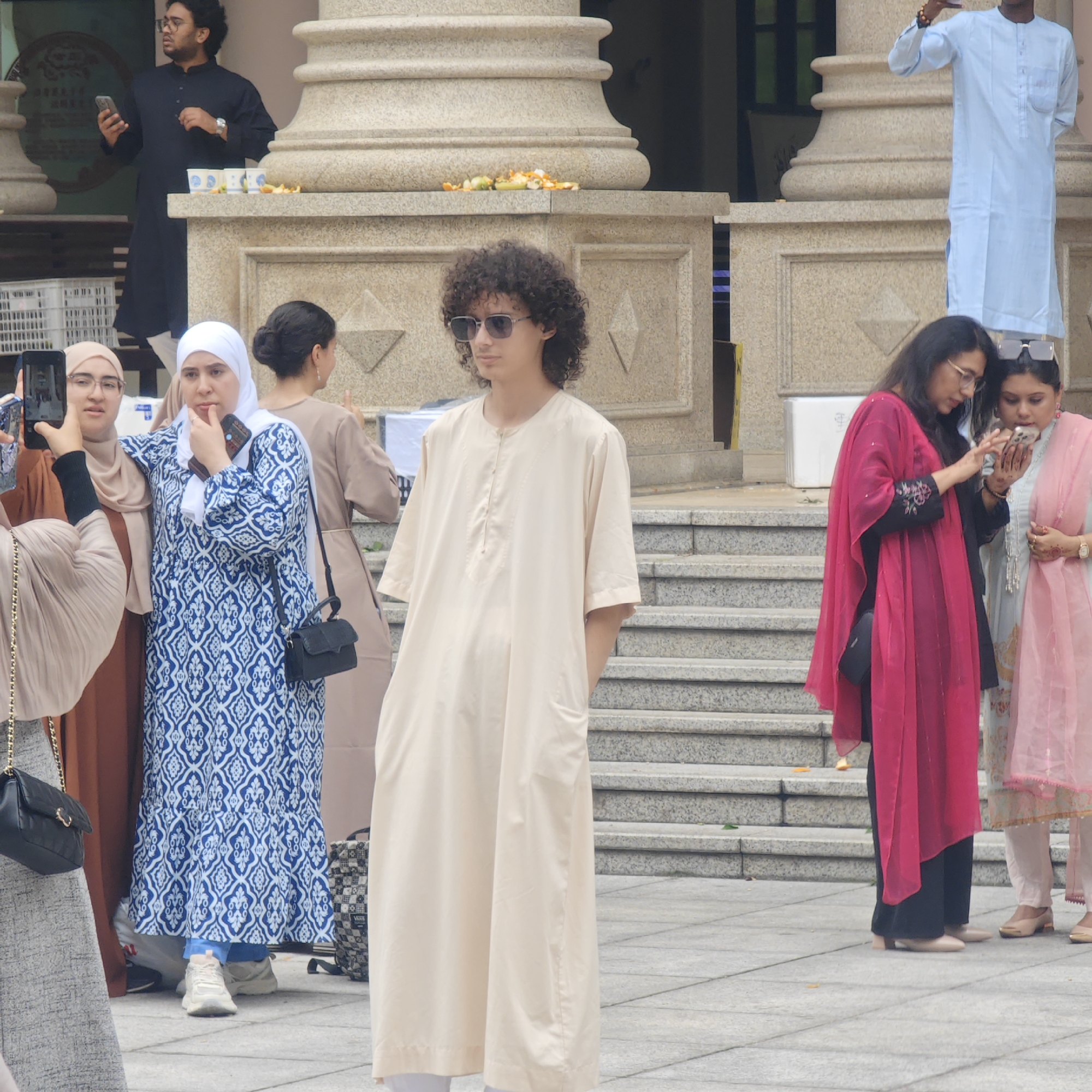 The image depicts a group of people standing in front of a building with large pillars. The individuals are dressed in a variety of clothing, some appearing to be traditional or religious garb. Several people are using mobile phones. The setting appears to be urban, with a building featuring architectural details such as pillars and decorative elements. It's daytime, and the overall impression suggests a public gathering or event. There are fruits on the platform in front of the pillars.
The location is Jiulongpo District, China.
