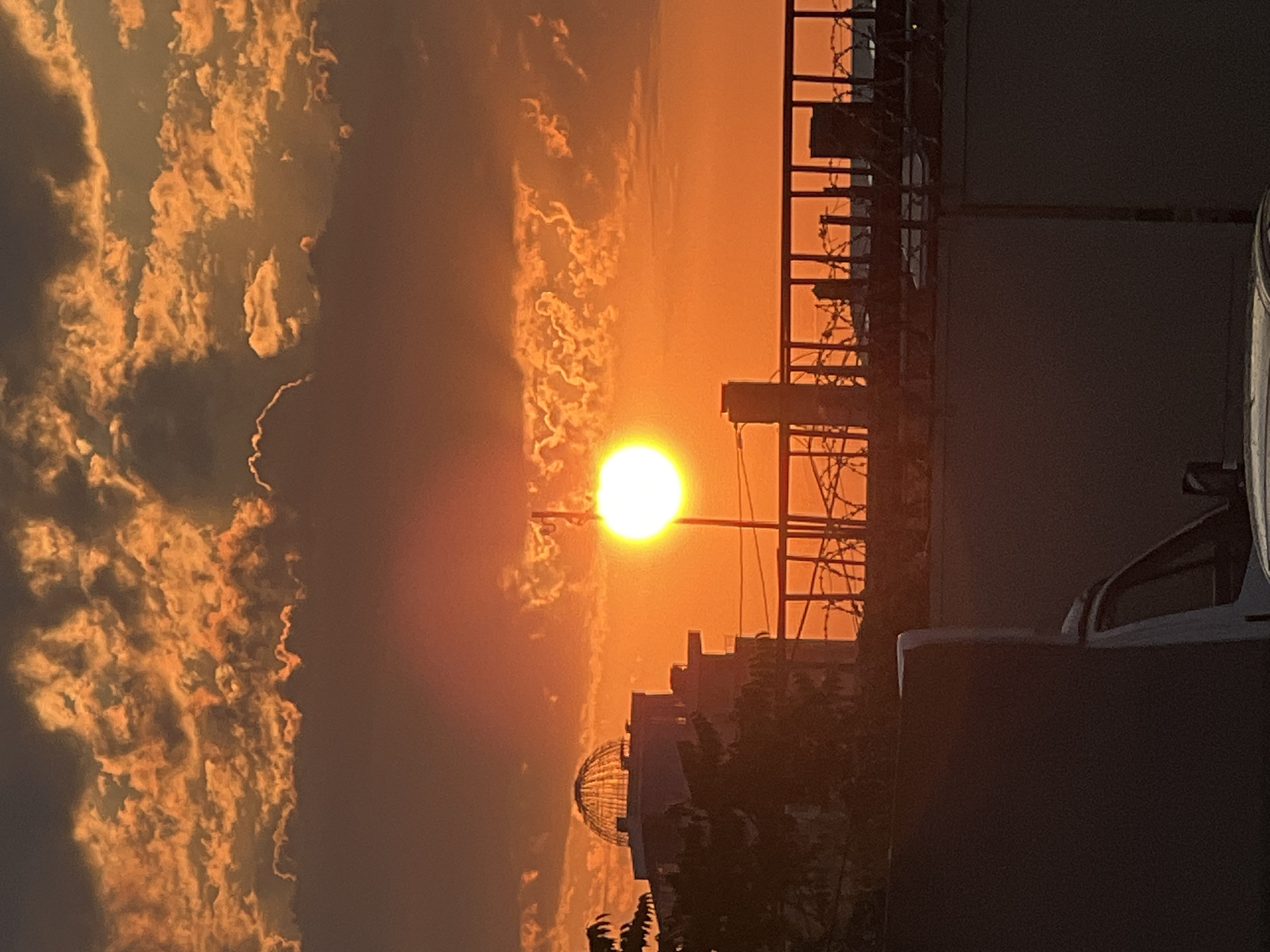 This is a vertical, medium-close shot of a sunset in Krasnodar, Russia. The sun is a bright, yellow orb low in the sky, casting an orange glow across the scene. Wispy, golden clouds are scattered throughout the sky, some appearing textured and almost like cotton candy.
In the foreground, to the right, is a dark, silhouetted structure that appears to be a building or part of a building. A portion of a vehicle, possibly a white car, is visible on the far right, its shape partially obscured by the darkness and glare. Further left, and lower in the frame, is a darker silhouette of trees and what looks like the top of a building with a domed structure.
To the left of the sun, a tall, dark, industrial-looking structure with horizontal and vertical bars and what appears to be barbed wire at the top is silhouetted against the bright sky. This structure creates a stark contrast with the soft, warm colors of the sunset.
The overall impression is one of a dramatic and beautiful sunset, with the industrial elements adding a touch of urban realism to the natural spectacle. The time of day is clearly sunset due to the low angle of the sun and the warm, diffused light. The weather appears clear, allowing for unobstructed views of the sunset and clouds. The scene evokes a sense of calm and wonder associated with sunsets. There is no visible text in the image.