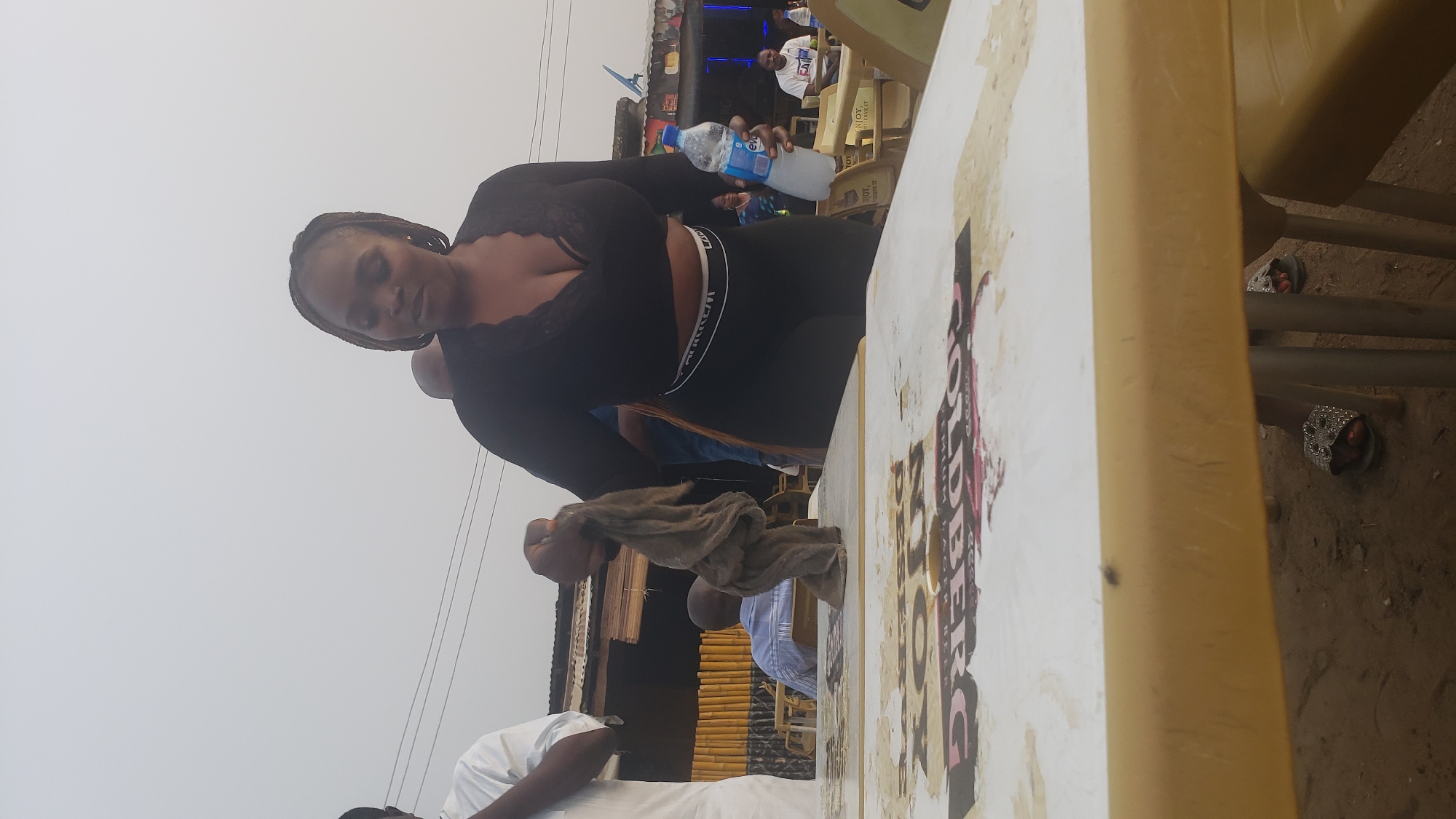The scene shows a woman holding a water bottle, possibly at an outdoor event in Narayi, Nigeria. The woman has braided hair and is wearing a black long-sleeved top and black leggings with a white band. Behind her are other people, chairs, and what appears to be a structure with bamboo-like siding. A table with peeling advertisements on it is in the foreground. The ground around the table is dirt, and there is a pair of sandals under the chair next to the table. Overhead are electrical wires. The sky is overcast.