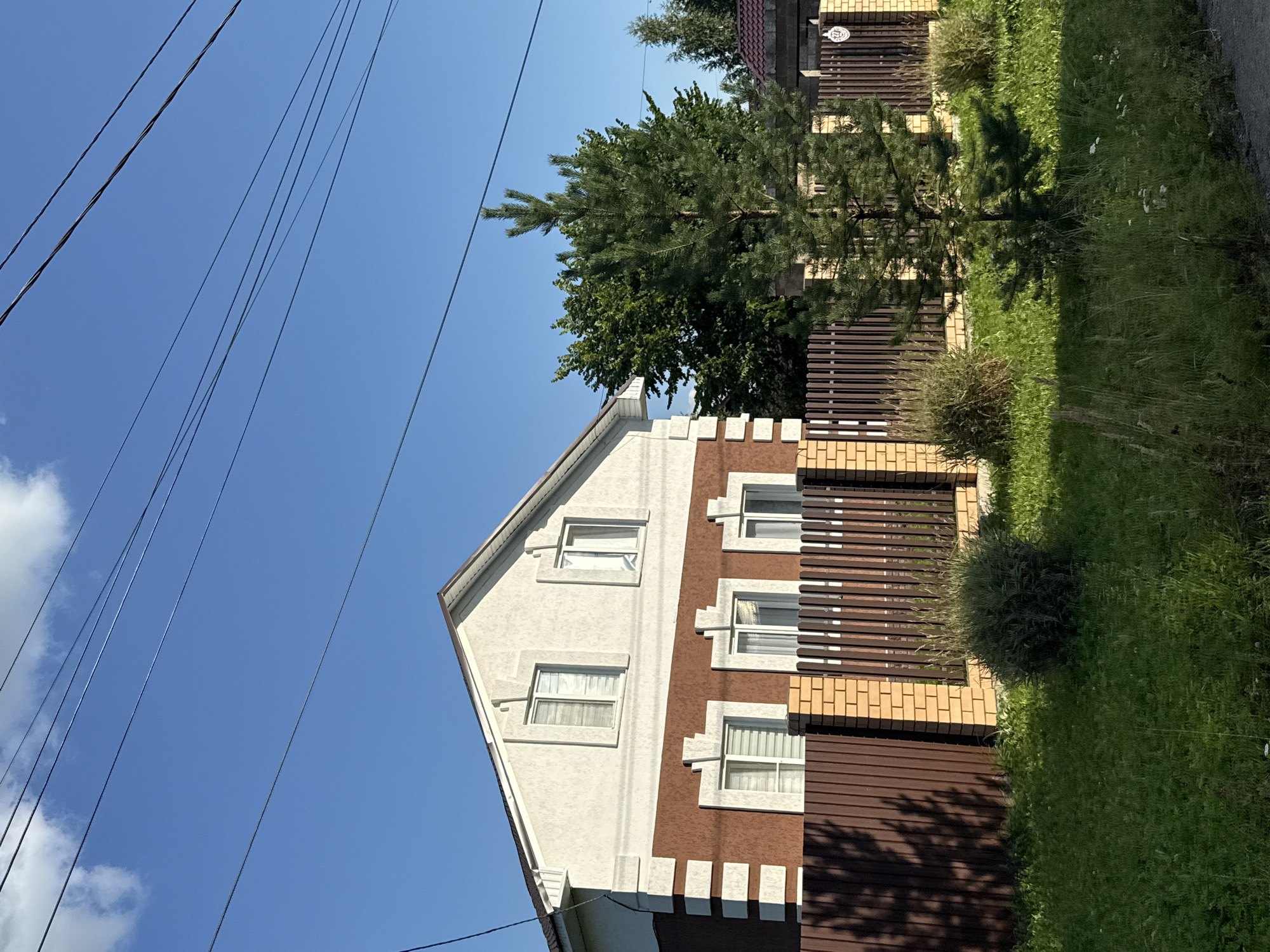 This is a photograph of a two-story house with a brown and white facade, located on a sunny day in the city of Nagayevo, Russia. The house has a pitched roof and several windows on both floors, some with light-colored blinds. A wooden and brick fence surrounds the property, with tall, dark brown vertical planks and lighter colored brick posts. In front of the fence, there are several green bushes and a young pine tree. The ground is covered with green grass, and in the background, there are more trees and a clear blue sky with some clouds and overhead power lines. There are no people or animals visible in the image.