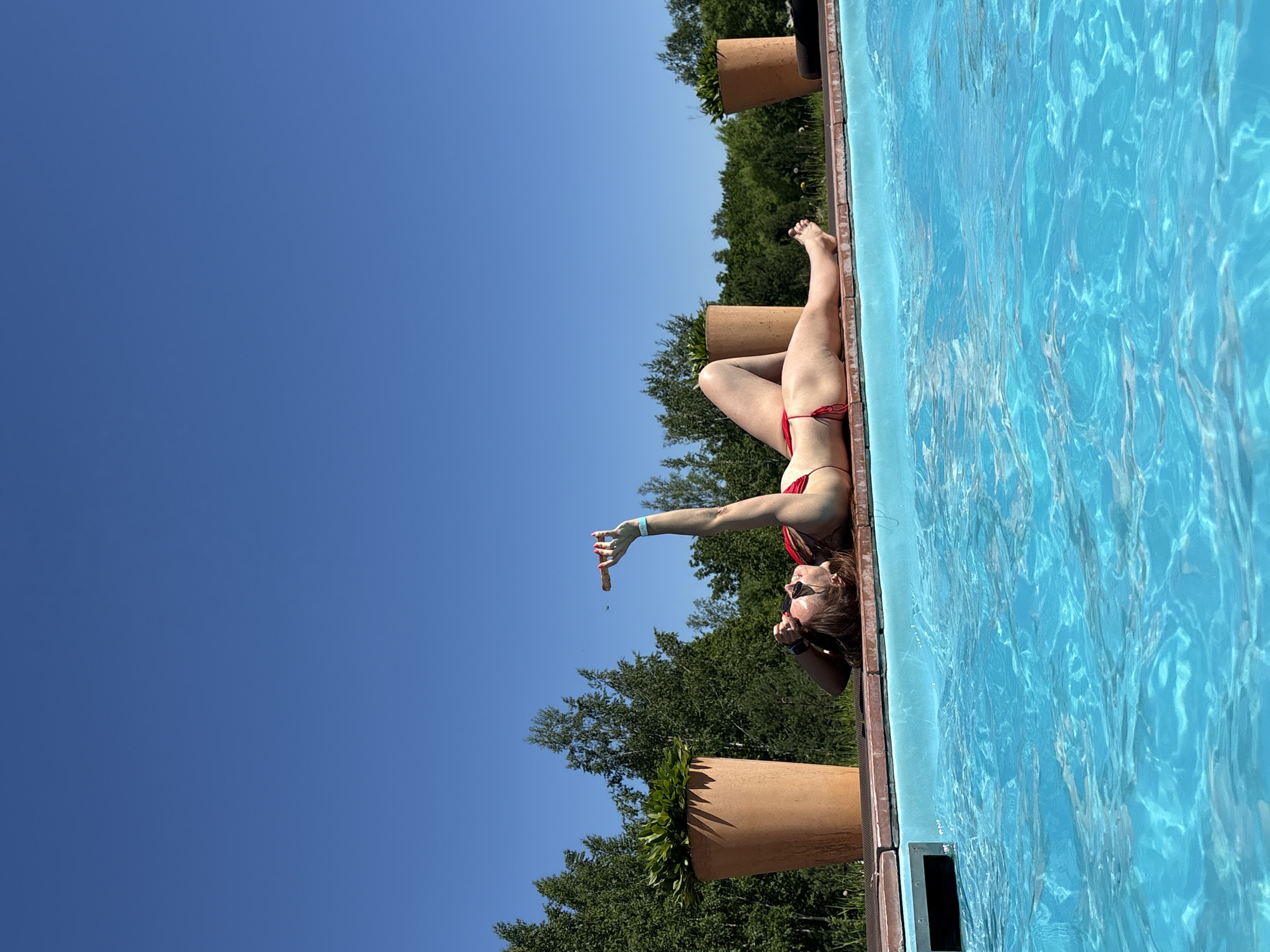 A woman is lying on her back on the edge of a swimming pool on a sunny day. She is wearing a red bikini and sunglasses. Her right arm is raised, and she is holding a cigar. The pool is filled with bright blue water. In the background, there are trees and two large terracotta pots with plants. The sky is clear and blue. The location is identified as Birobidzhan, Russia.