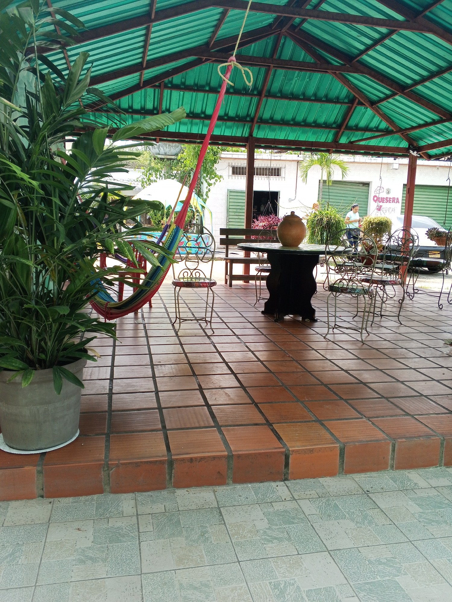 Outside a building labeled "QUESERA," in Tinaquillo, Venezuela, a covered patio features a brown-tiled floor. A large potted plant is in the foreground. A colorful hammock hangs nearby. Wrought iron chairs surround a dark table with a large clay pot on it. A person stands near the edge of the patio.