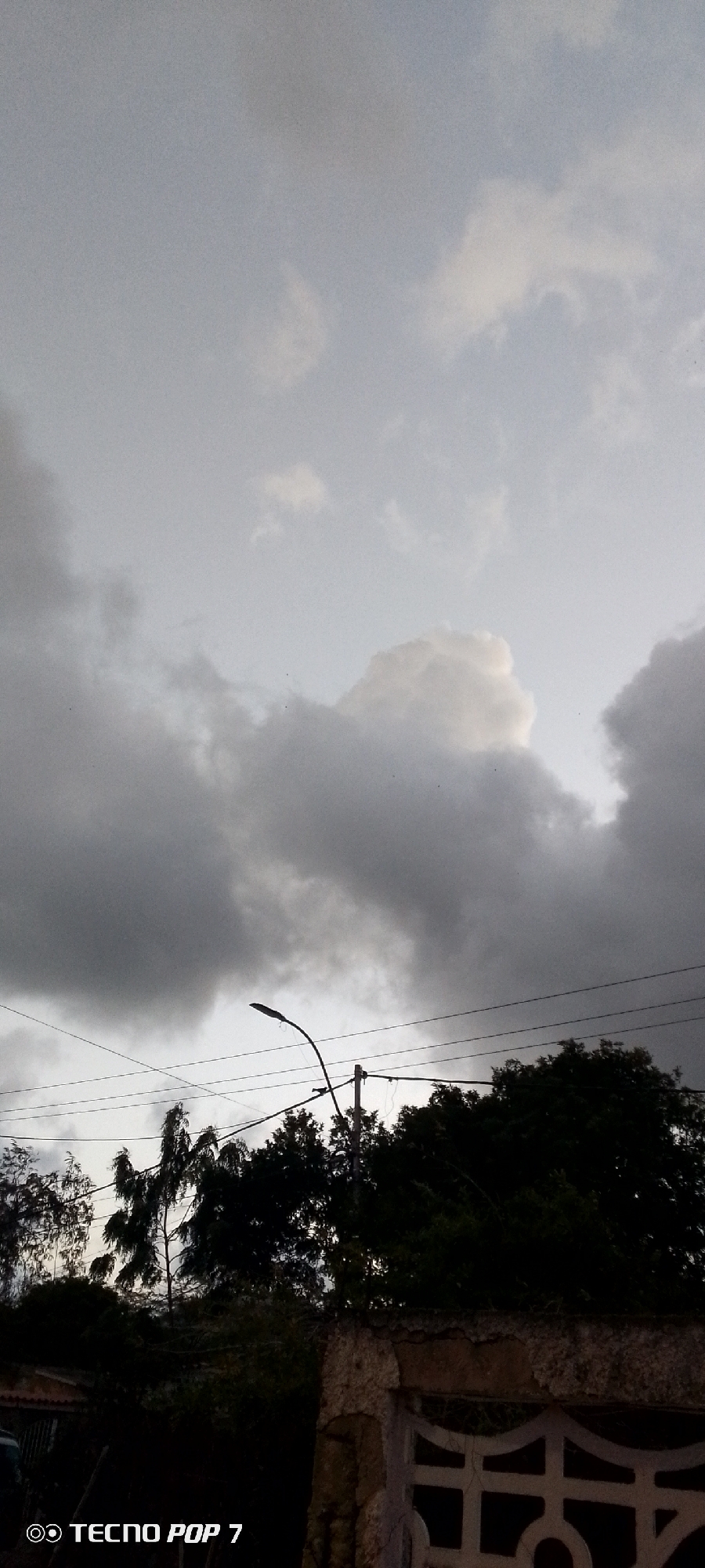 The image shows a cloudy sky above the city of Altagracia, Venezuela. Below the sky is a line of trees and a street light. There is a wall in the foreground with an ornamental design. The TECNO POP 7 watermark is visible in the bottom left corner of the image.