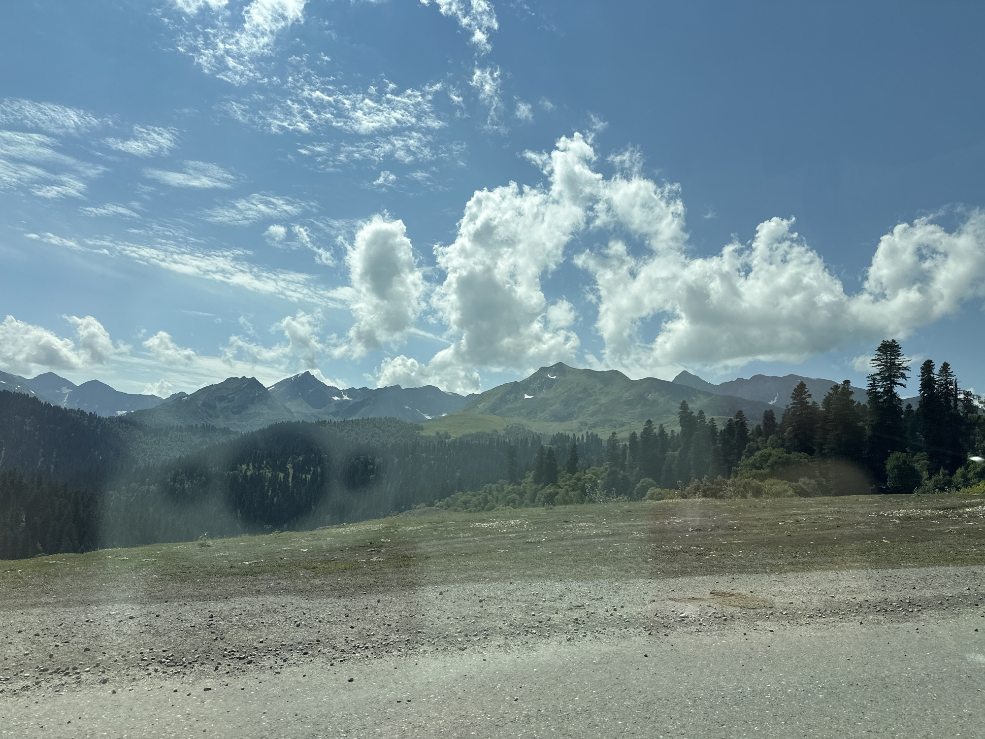 The image depicts a vast, mountainous landscape under a partly cloudy blue sky. The foreground shows a rough, gravelly road with some patches of sparse grass. Beyond the road, a gently sloping meadow is visible, transitioning into a dense forest of coniferous trees that covers the lower slopes of the mountains. The mountains themselves are layered, with some peaks showing signs of snow. The sky is filled with a mix of wispy cirrus clouds and larger cumulus formations. The scene appears to be captured from a moving vehicle, as suggested by a slight blur and a reflective surface in the foreground. The location is identified as Lunnaya Polyana, Russia.