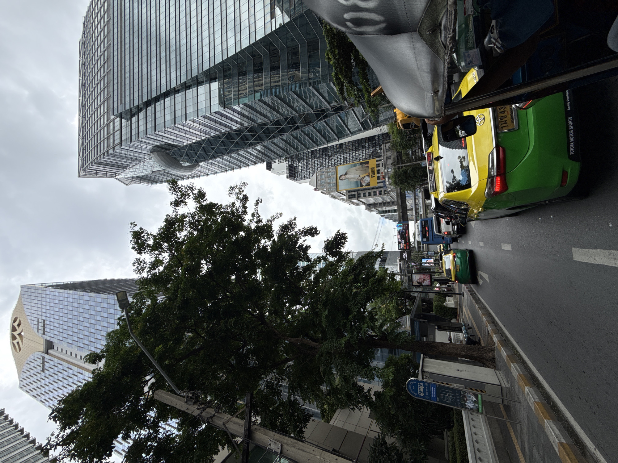The image captures a street scene in Bangkok, Thailand, on an overcast day. Tall, modern glass buildings dominate the skyline, with one building on the left featuring a distinct golden, geometric crown. A large, mature tree with dense foliage stands in the foreground on the left, partially obscuring a concrete utility pole and a bus stop sign. The street is congested with traffic, including a yellow and green taxi, a tuk-tuk in the immediate foreground, and other vehicles. A large billboard featuring a person in a suit is visible on the side of a building in the mid-ground. The road surface is asphalt, marked with white lines. The overall impression is of a busy urban environment with a mix of contemporary architecture and lush greenery.
