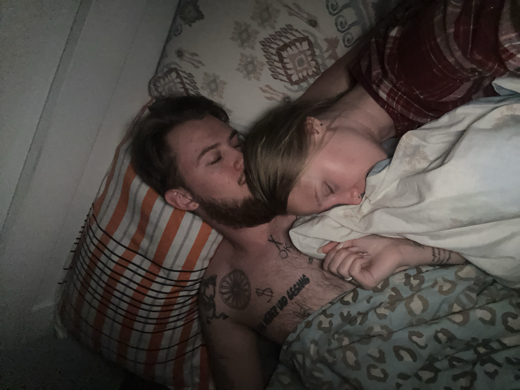 A man and a woman are lying in bed. The man is on his side, facing the woman, and appears to be kissing her on the cheek. Both have their eyes closed. The man has a beard and tattoos on his chest and upper arm. The woman is partially covered by a blanket with a white sheet and a blue leopard-print duvet cover. The bed has two pillows with different patterns. The background features a white wall and a white and orange pillow with stripes. The location is not discernible from this image.
