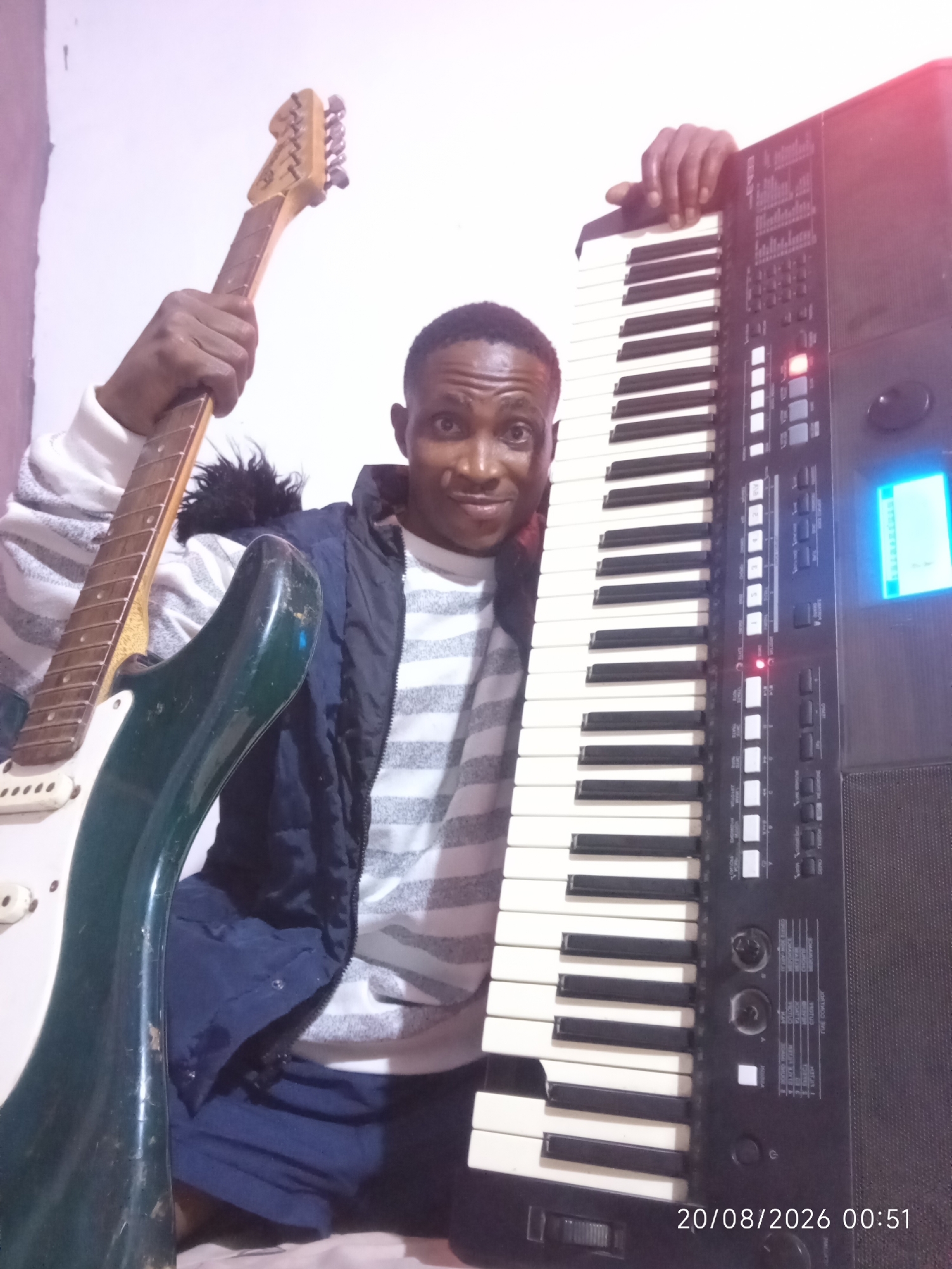 A young Black male, likely in his late teens or early twenties, stands in what appears to be a residential setting, possibly in Adankolo, Nigeria. He is holding a black and green electric guitar in his left hand, with the neck extending upwards. His right hand rests on a black keyboard, which dominates the right side of the frame. The male is wearing a horizontally striped long-sleeved shirt under a dark blue puffer vest. He is looking directly at the camera with a surprised or excited expression, his eyes wide open. The background is a plain, light-colored wall. The lighting suggests an indoor environment, possibly with a flash due to the stark contrast and slight overexposure in some areas. A timestamp in the bottom right corner reads "20/08/2026 00:51".