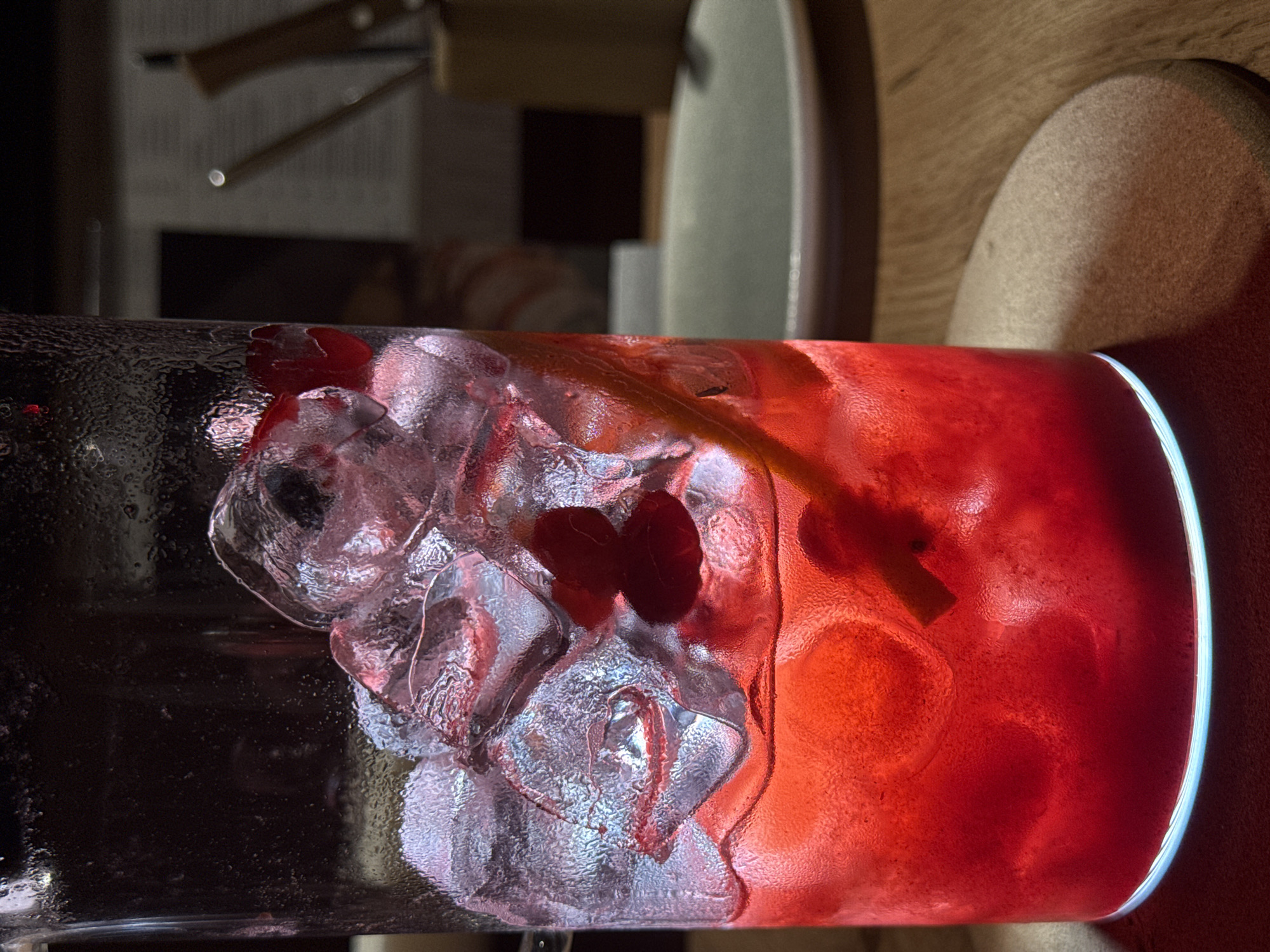 A close-up, horizontally-oriented image shows a tall glass filled with ice cubes and a red liquid, possibly a cocktail or juice. The glass is positioned horizontally, with the left portion of the glass filled with ice, some red fruits or berries visible among the ice. The right portion is filled with the red liquid, also containing some solid ingredients like fruit slices. The exterior of the glass has condensation droplets.
The background is out of focus. A light wooden surface is partially visible behind the glass, and in the top left corner, there is a glimpse of a chair or table leg. Part of a person's body (thigh area) is in the far right of the image.
The location is not discernable as Chaikovsky, Russia from visual cues.
