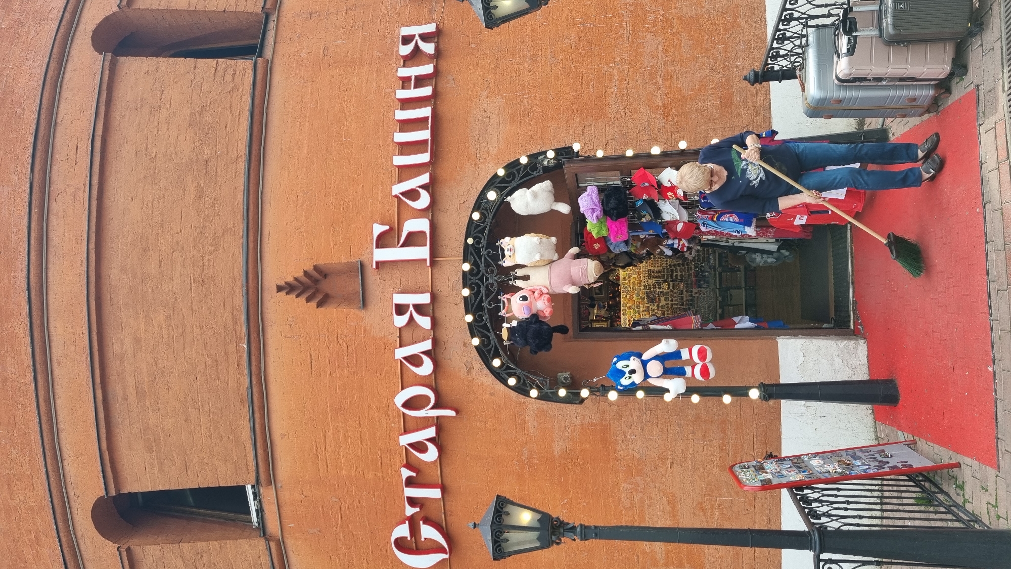 A woman with short blonde hair and glasses is sweeping a red carpeted area in front of a store with a large arched entrance. The store is adorned with string lights and hanging plush toys, including a Sonic the Hedgehog toy. The store's name, "Старая Башня," is written in large white letters with red trim above the entrance. The building is constructed of orange brick. Several suitcases are visible to the right of the entrance. A lamppost stands to the left of the entrance. A red sign with photos is propped up near a black metal fence.