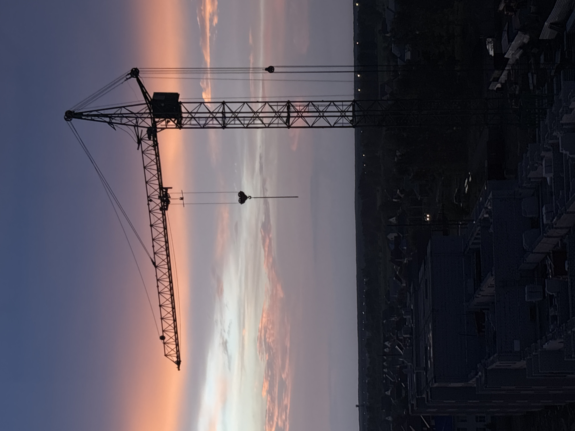 The image depicts a construction site at dusk in Novoaltaysk, Russia. A large tower crane dominates the frame, silhouetted against a twilight sky transitioning from deep blue to hues of orange and pink from the setting sun. Below the crane, the dark shapes of a partially constructed building are visible in the foreground, constructed with grey bricks. Further in the distance, a residential area with scattered lights can be seen, nestled amongst dark trees. The overall scene conveys a sense of ongoing development and the quiet transition from day to night in a Russian city. There are no people visible in the image.