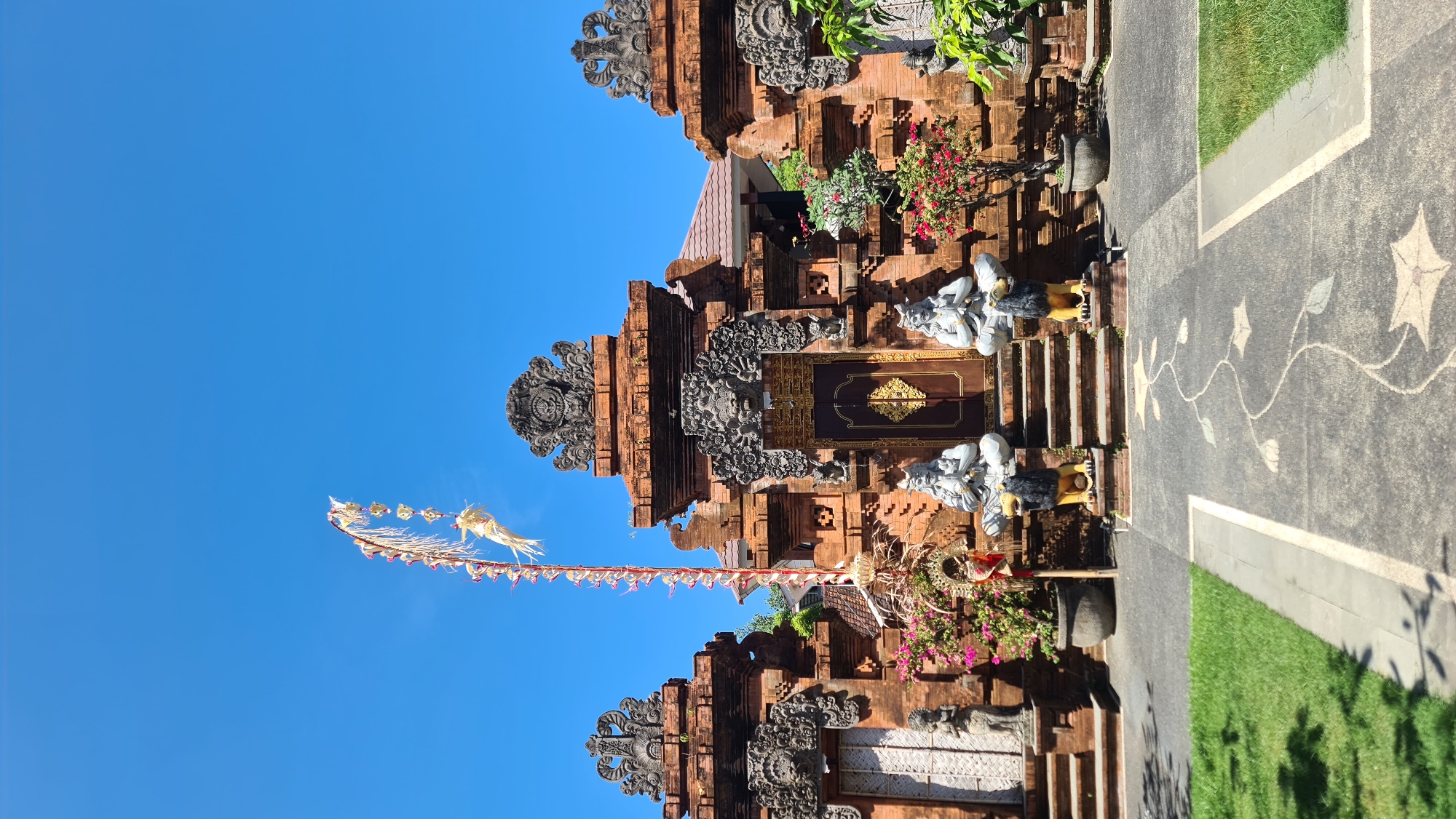 The image shows the entrance to a temple in Denpasar, Indonesia, under a clear blue sky. The temple is constructed of reddish-brown brick and features intricate stone carvings above the doorways. Two large statues of seated figures, each with a lion-like creature at its base, flank the central entrance. A tall, decorative pole adorned with natural materials stands to the left of the entrance. Pink flowers bloom in pots near the entrance, and there is a well-maintained green lawn in the foreground. The path leading to the temple is paved with dark grey stones decorated with a lighter grey floral motif.