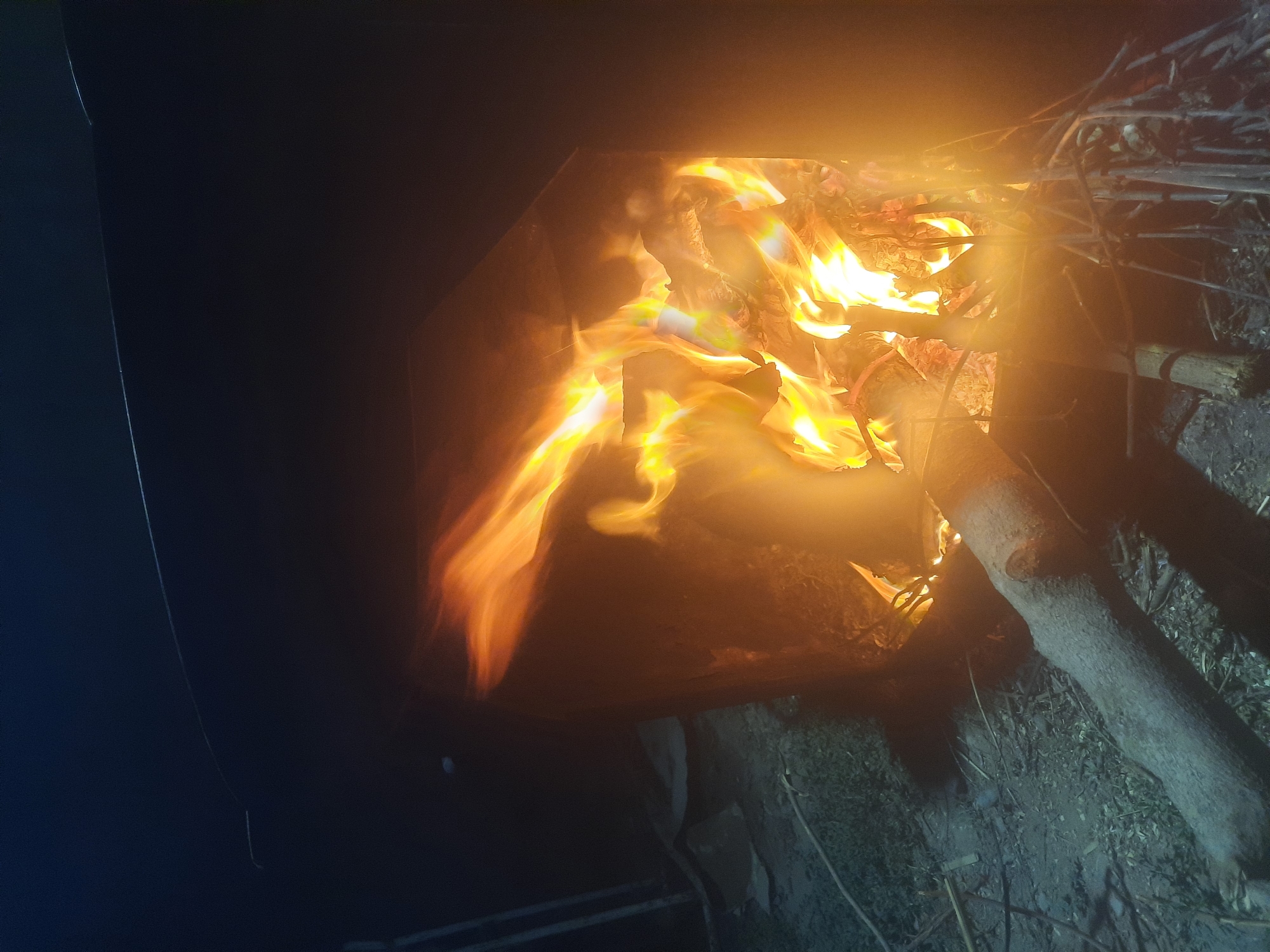 The image depicts an outdoor scene at night, likely in Восеабад, Tajikistan, featuring a bonfire. The fire burns brightly within a dark, possibly metal, enclosure, casting a warm glow on the surrounding area. Flames of orange and yellow dance upwards, illuminating logs and smaller branches within the fire pit. In the foreground, a thick log lies horizontally on the ground, with smaller sticks and debris scattered around it. The background is dark, with hints of vegetation or ground cover visible in the dim light. The overall impression is one of a rustic, nighttime setting centered around a crackling fire.