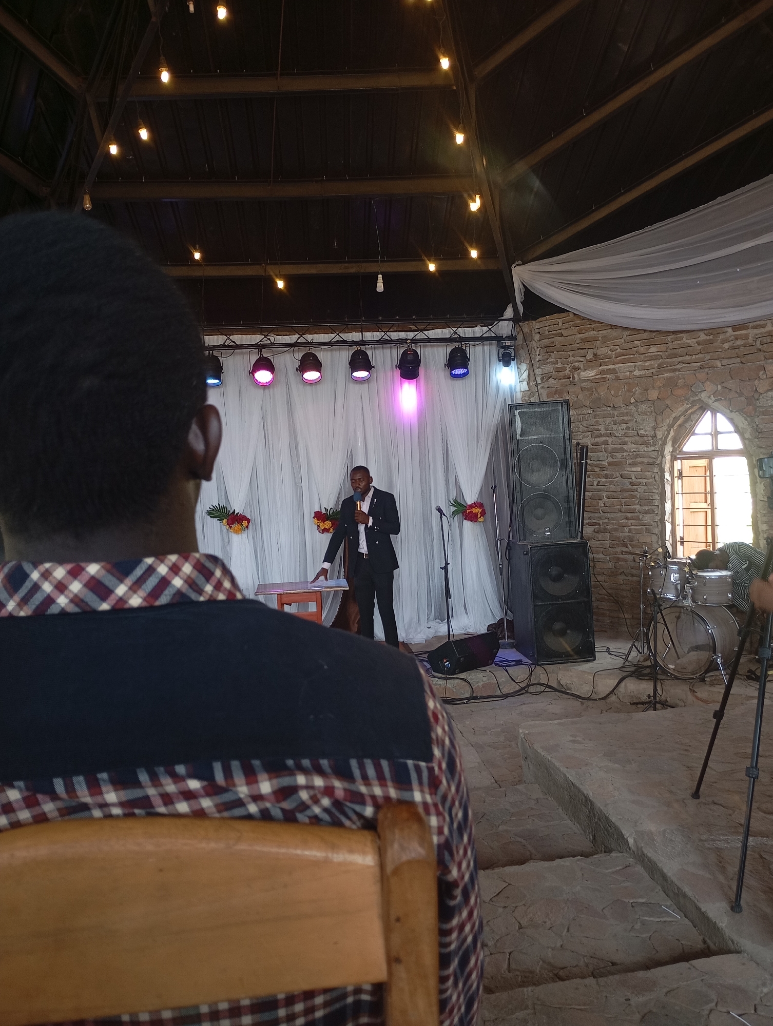 A medium shot captures a man speaking on a stage in what appears to be a church or event hall in Uke, Nigeria. The speaker, dressed in a dark suit, stands behind a small wooden podium and holds a microphone. Behind him is a white curtain backdrop lit by stage lights. To his right, large speakers flank the stage, with a drum set positioned near a window.
In the foreground, the back of another man's head is visible, seated in a wooden chair, suggesting he is part of the audience. The floor is made of stone, and the hall's ceiling is visible, featuring exposed beams and hanging lights. The setting indicates an indoor event, likely a religious service or a public speaking engagement.
