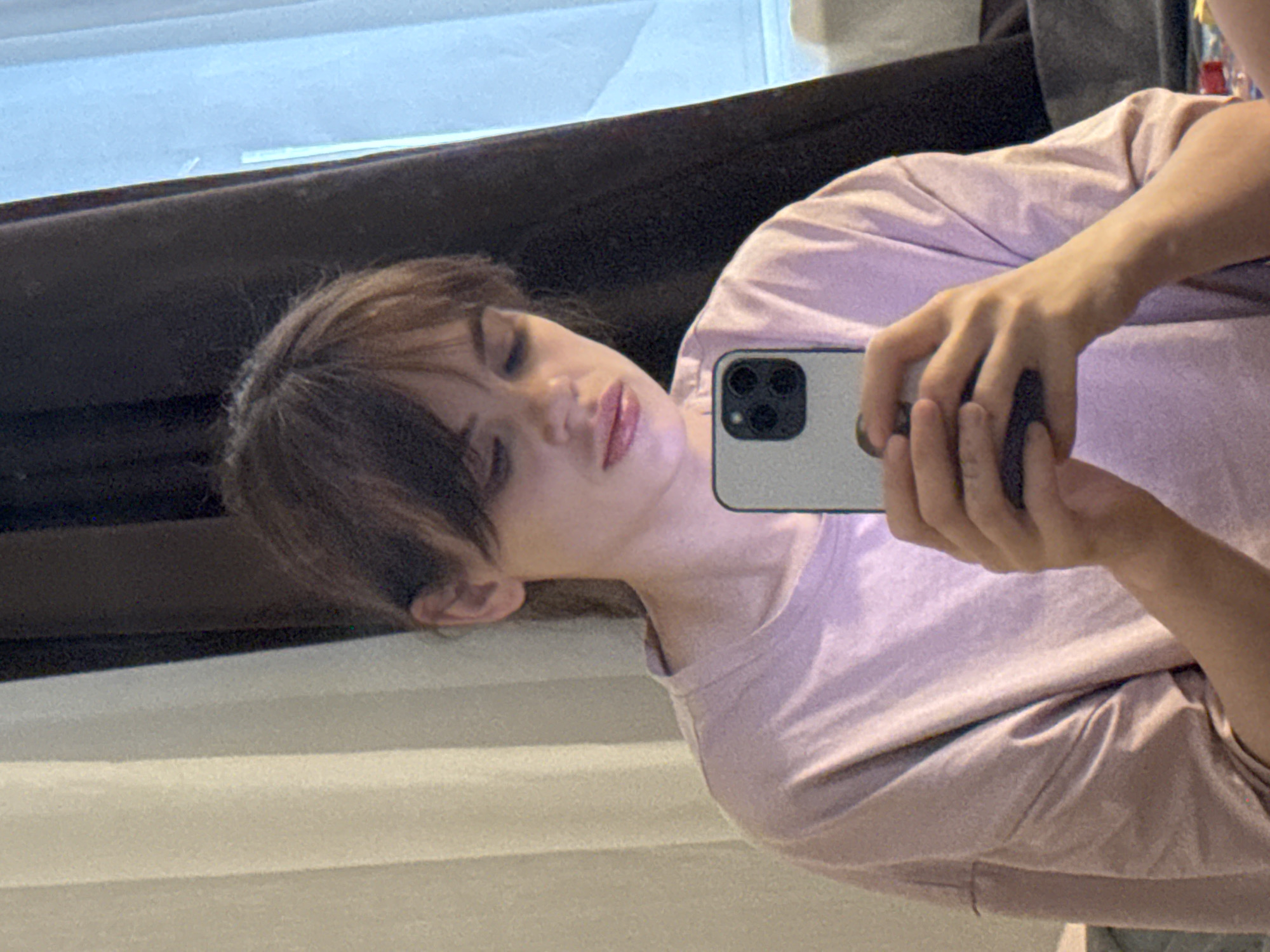 A person is taking a selfie with a phone. The person has brown hair with bangs. The person is wearing a light pink shirt. The background includes a window and part of what appears to be a dark-colored headboard or curtain. There is not enough information to determine the location of the photo in Rostov-on-Don, Russia.