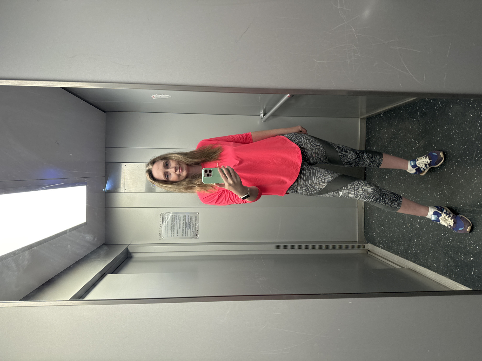 A woman is taking a selfie in an elevator. She has blonde hair and is wearing a bright pink long-sleeved shirt, patterned leggings, and blue and white sneakers. She is holding a green smartphone. The elevator has gray metal walls, a speckled gray floor, and a bright light fixture. A sign is mounted on the wall. The location is possibly Chaikovsky, Russia, based on the request.