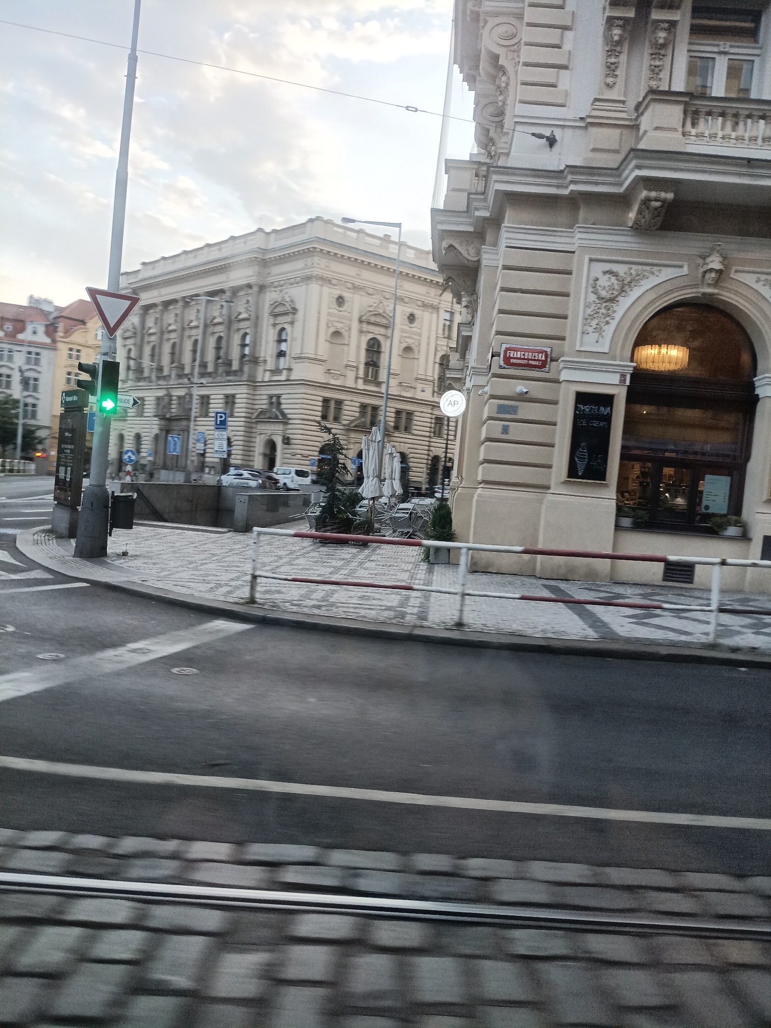 The image captures a street scene in Prague, Czechia. A traffic light shows a green arrow, indicating a permitted turn. In the foreground, a cobblestone street and a road with white lane markings are visible. Beyond the road, a pedestrian crossing leads to a sidewalk with outdoor seating for a cafe or restaurant. The architecture of the buildings suggests a European city, with ornate facades and classical elements. A sign with the name "FRANCCOISKA" is displayed on one of the buildings. The sky is overcast, with soft clouds. The overall impression is a typical urban street in a historic European city.
