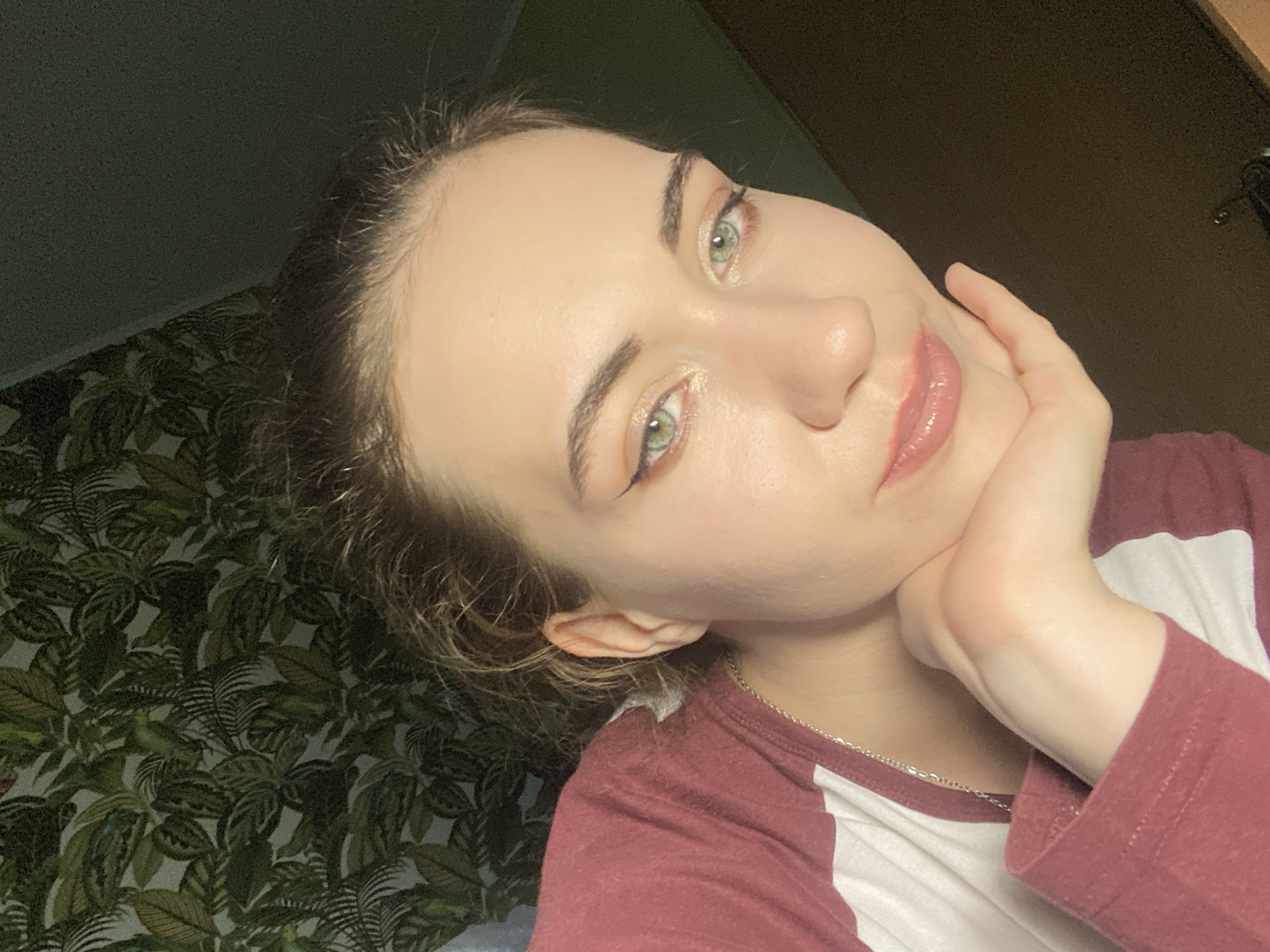 This is a close-up, head-and-shoulders portrait of a young woman indoors. She is resting her right hand on her chin, looking towards the viewer with a soft expression. Her eyes are green and appear to have makeup on, including eyeliner and eyeshadow. Her lips are glossy and a shade of pink. She has light-colored hair pulled back, with some strands loose around her face. She is wearing a maroon and white baseball-style t-shirt.
The background consists of a dark wall with patterned wallpaper featuring green leaves. A wooden surface is visible in the upper right corner. The lighting suggests it is likely evening or indoors with artificial light. No visible text is present in the image. The location context is Zavodoukovsk, Russia.