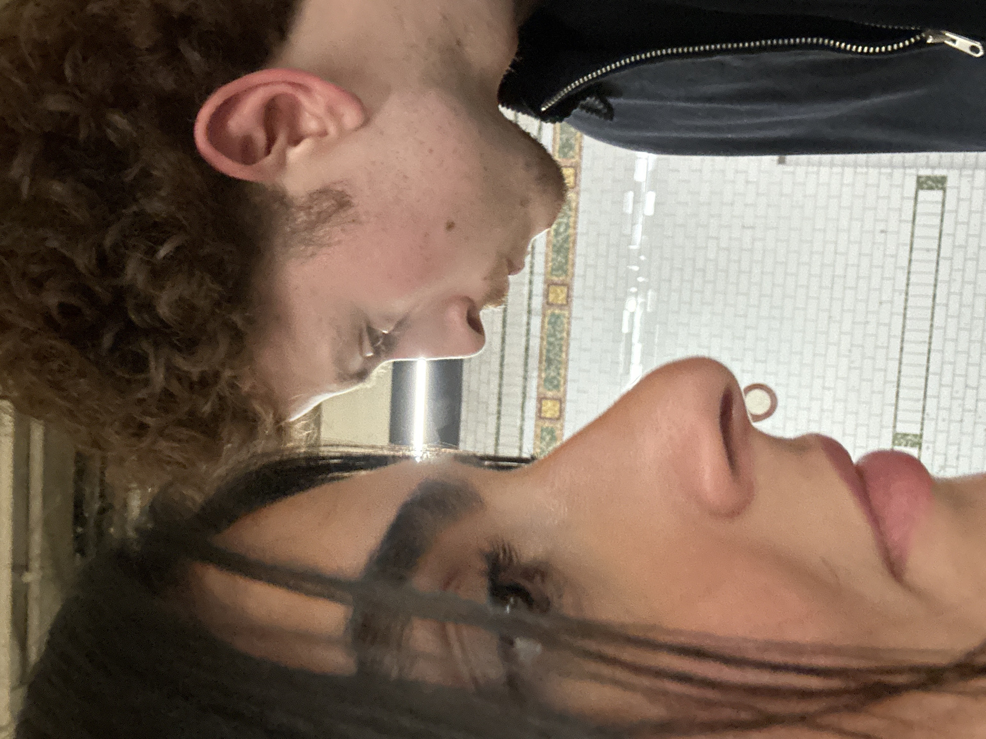 The image depicts two individuals in close proximity within a New York City subway station, United States of America. The background consists of white rectangular subway tiles with dark grout lines and a decorative horizontal band of green and yellow patterned mosaic tiles. Overhead artificial lighting is visible.
In the foreground on the left, a person with long, dark hair is shown in profile from the nose downward. A small C-shaped septum piercing is visible. The person's left eye is partially visible, appearing to gaze towards the second individual.
In the background on the right, a second person with light brown, curly hair and light facial hair (mustache and stubble) is shown in profile. This individual wears a dark, possibly navy or black, zippered garment, with the silver zipper pull and teeth clearly visible on the right side of the frame.
The two individuals are positioned face-to-face, very close, with their profiles angled towards each other, suggesting an intimate interaction or conversation.