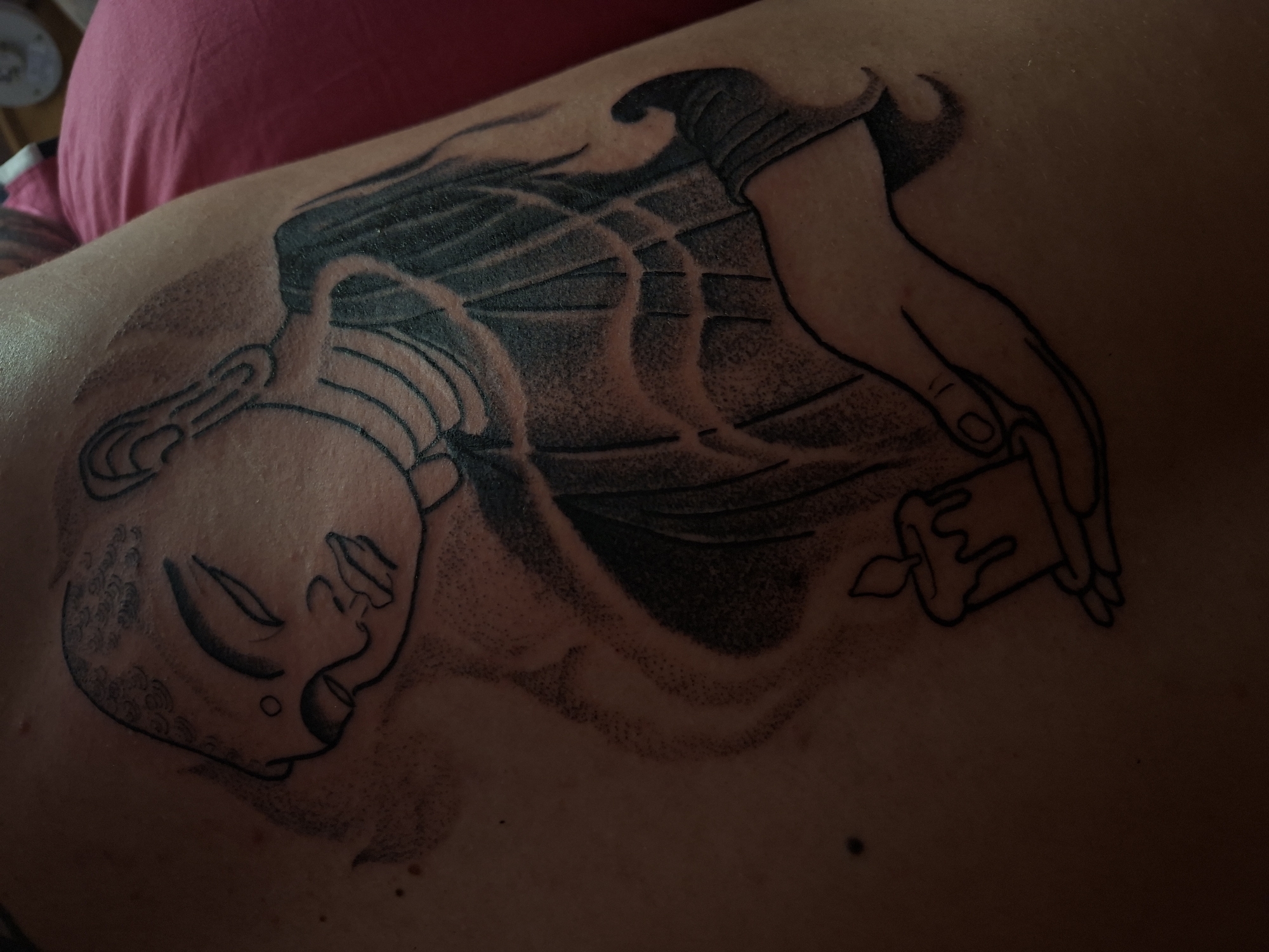 A close-up photograph shows a grayscale tattoo on a person's skin. The tattoo depicts a person lying down with their face angled to the left. A hand is shown holding a candle. The tattoo appears to be on the back or side of the torso of the person. A section of a pink shirt can be seen in the upper left of the photograph. The location is not discernable in the image.