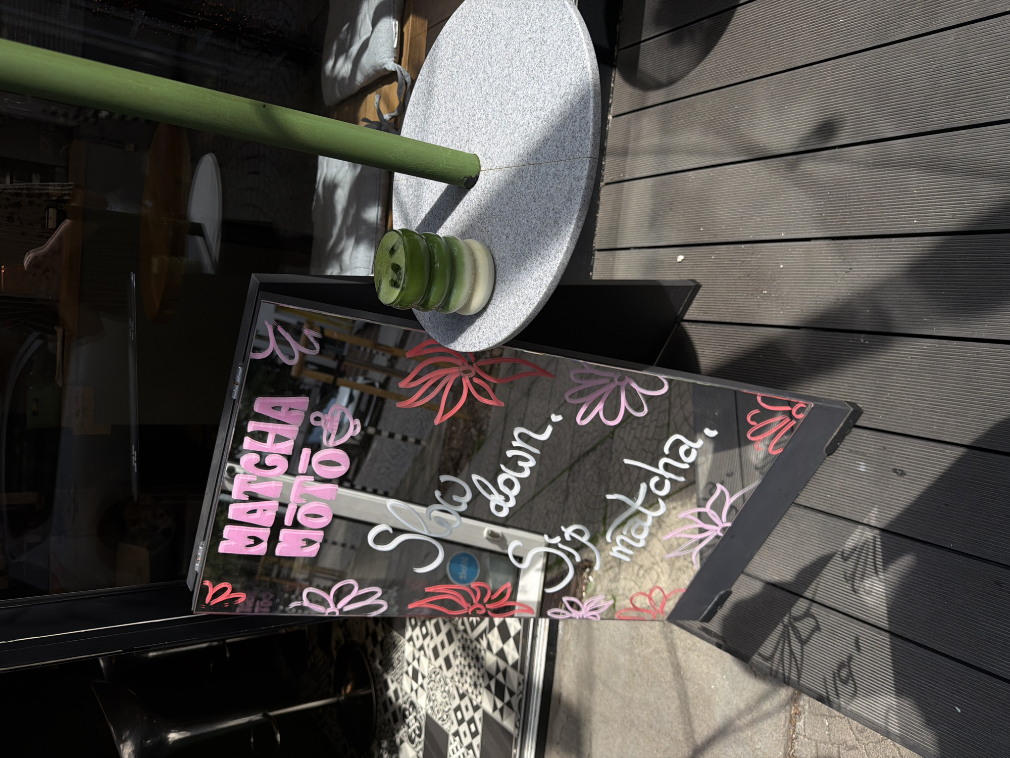The image depicts an outdoor scene in Sofia, Bulgaria, featuring a framed A-frame sign with text and decorations. The sign is placed on a dark wooden deck. On the sign, written in pink and white chalk, is the text "MATCHA MOTO" at the top, followed by "Slow down.", and "Sip matcha." along with floral drawings in pink and red. Behind the sign, a reflection in the glass shows part of a building interior with tables and chairs, and an exterior view with a street and possibly a car. To the right of the sign, a tall green pole stands vertically, originating from a speckled gray circular base on the wooden deck. On this base, there is a stack of three green cups or glasses, the top one filled with a green liquid, presumably matcha. The lighting suggests it is daytime, with shadows cast on the deck, indicating sunlight. The overall setting appears to be the entrance or outdoor seating area of a cafe or shop.