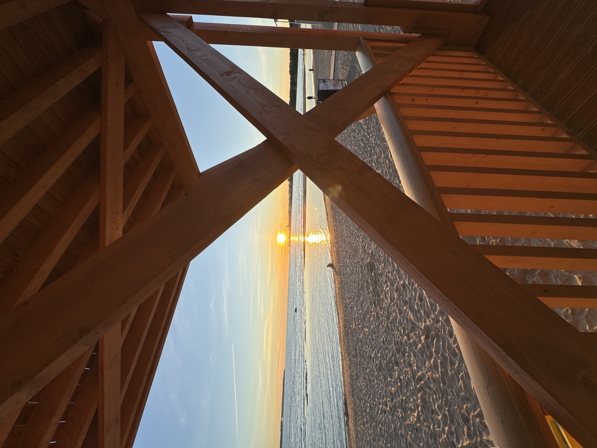 The image captures a sunset view of the Baltic Sea from a wooden structure on a beach in Tallinn, Estonia. The foreground is dominated by the warm tones of wooden beams and railings, forming a frame for the scene beyond. The sandy beach is visible, with gentle waves lapping at the shore. The sun, a bright orb on the horizon, casts a golden reflection across the water. In the distance, a silhouetted figure is visible on the beach, and a few other indistinct figures can be seen walking along the shoreline or on a boardwalk. The sky transitions from a clear blue near the top to hues of orange and yellow as it approaches the sunset. The overall atmosphere is serene and peaceful, characteristic of a late afternoon or early evening at the coast.
